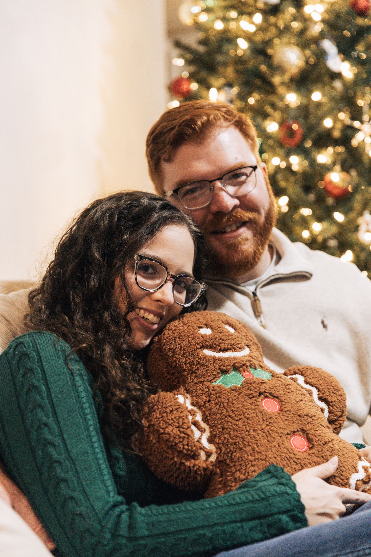 Couple shares intimate moment with gingerbread plush on couch near Christmas tree.