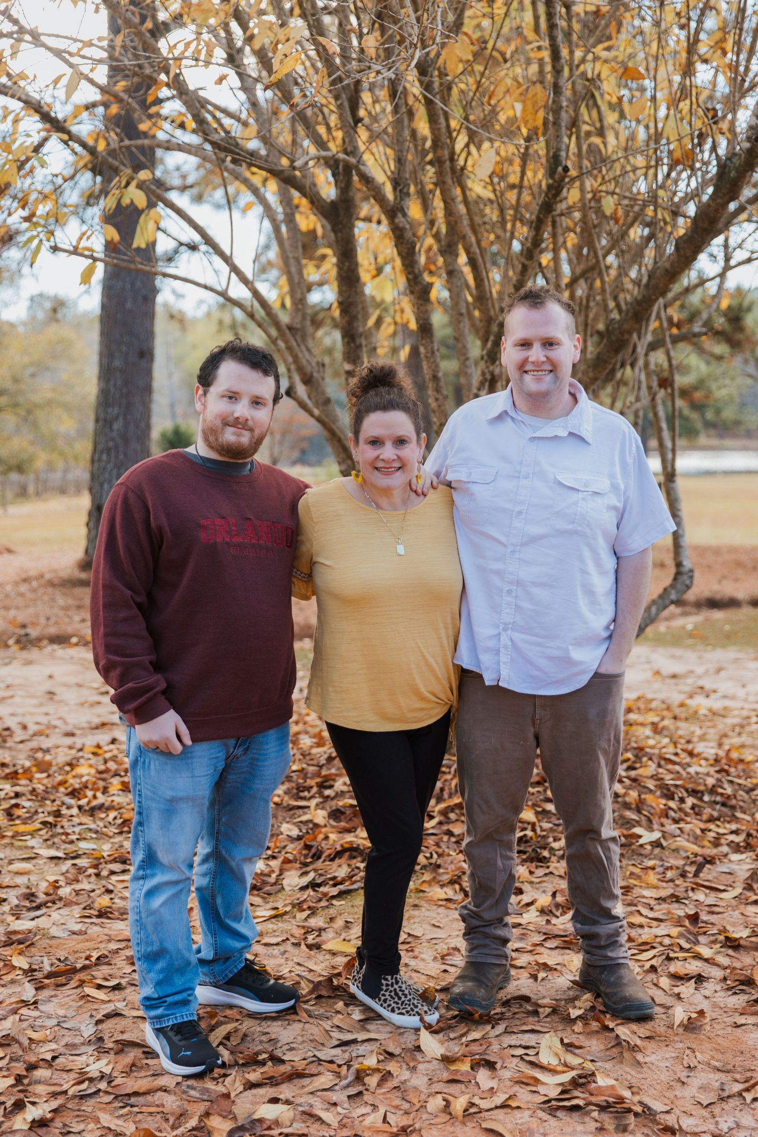 Three people posing together on fallen autumn leaves under trees in a park setting.