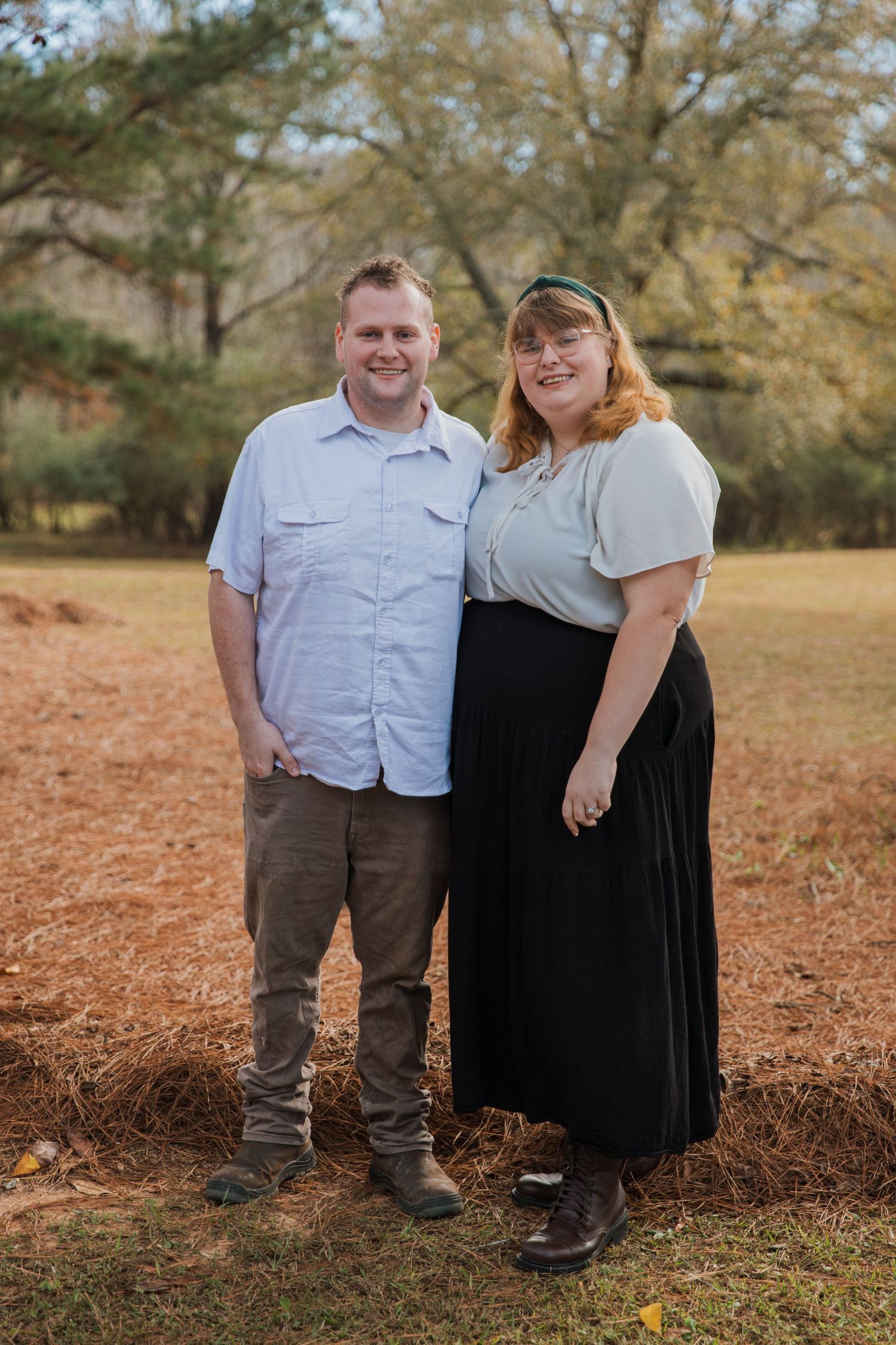 A couple stands together on red clay soil with autumn trees in the background during a casual outdoor photoshoot.