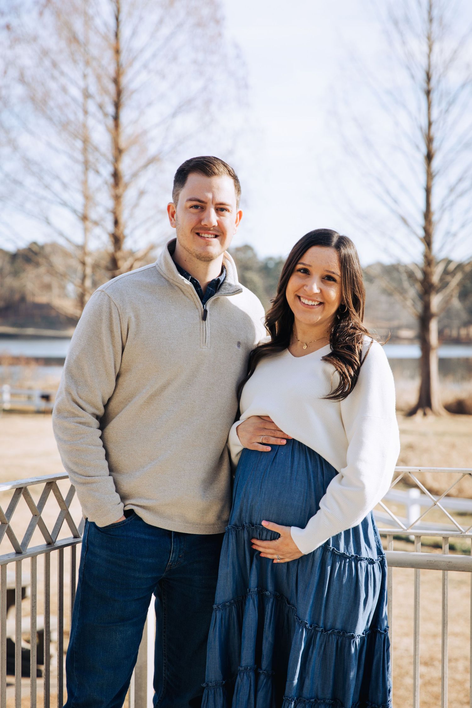 Couple in coordinating navy and beige outfits pose together outdoors in a winter field with bare trees in background.