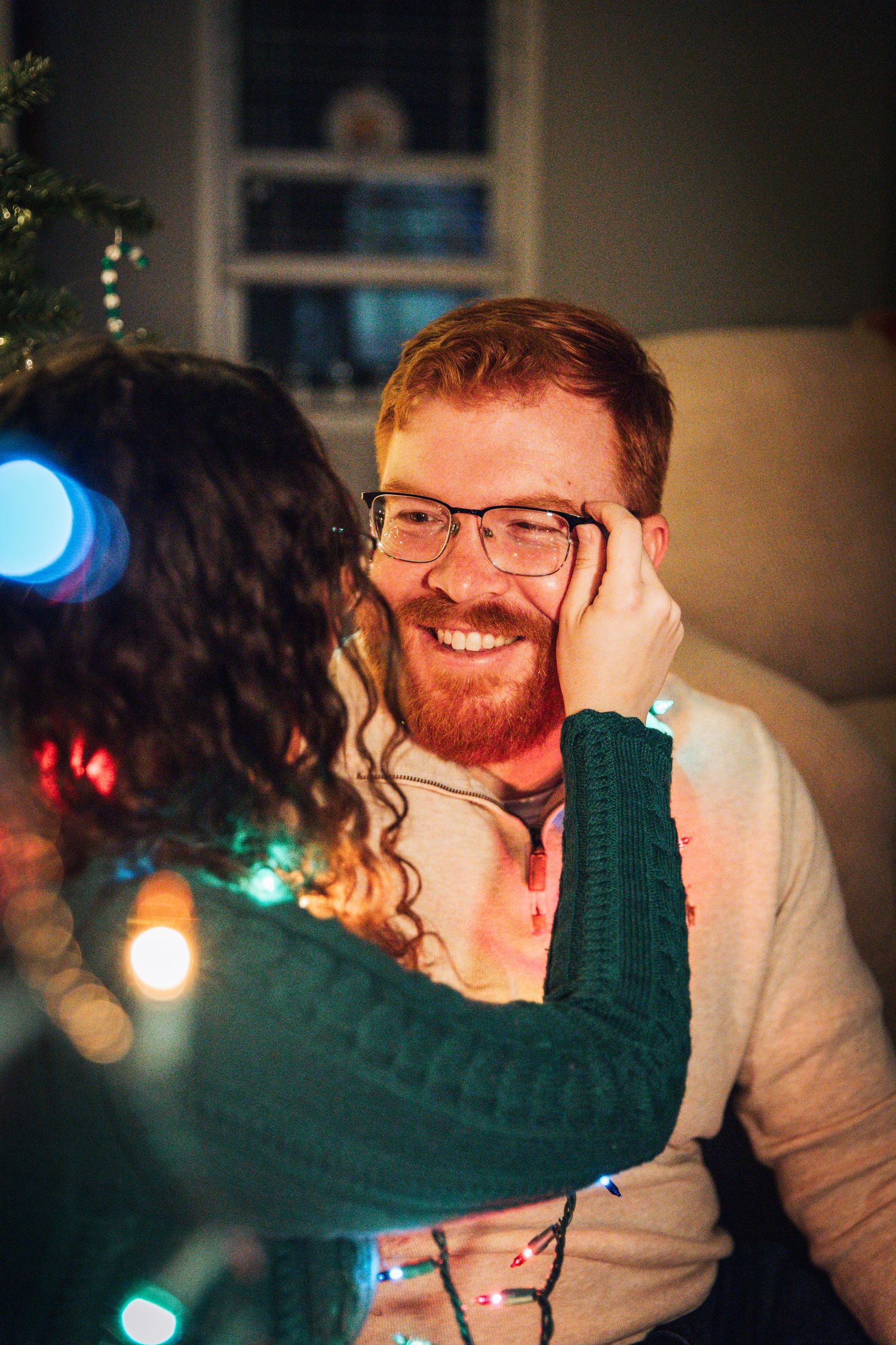 Couple sharing tender moments illuminated by colorful Christmas lights in living room setting.