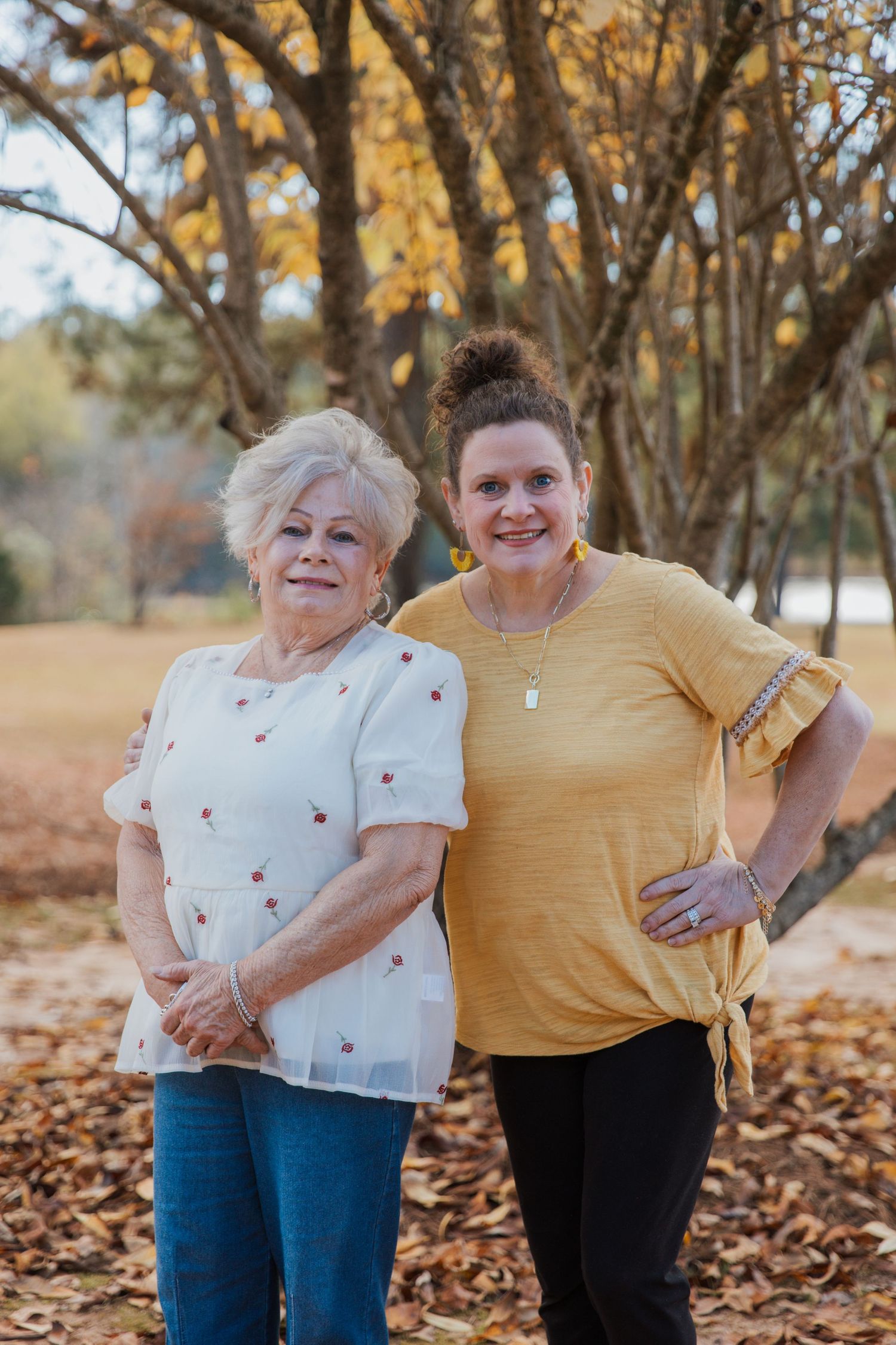 Two people share a warm embrace while standing among fallen autumn leaves and trees.
