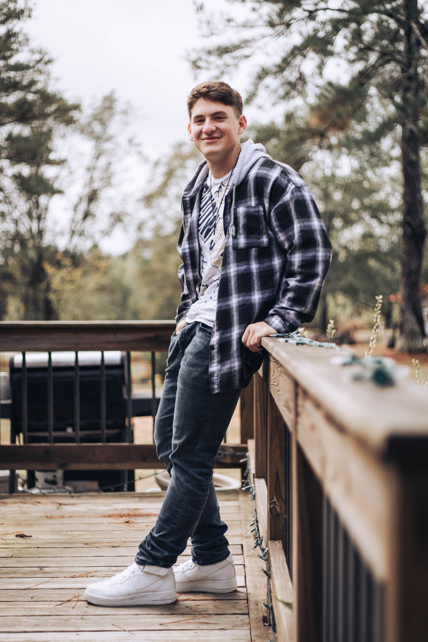 A person in a plaid flannel shirt and jeans poses casually on a wooden deck surrounded by pine trees.
