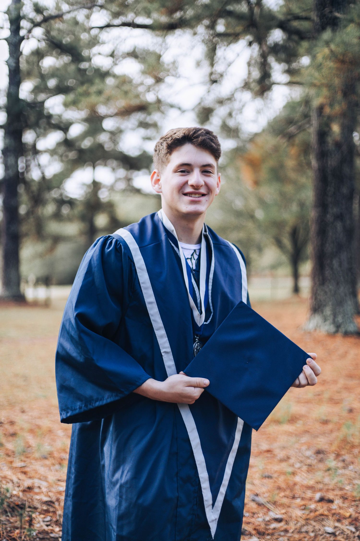A graduate in a navy blue graduation gown poses playfully in an autumn forest setting with fallen leaves.