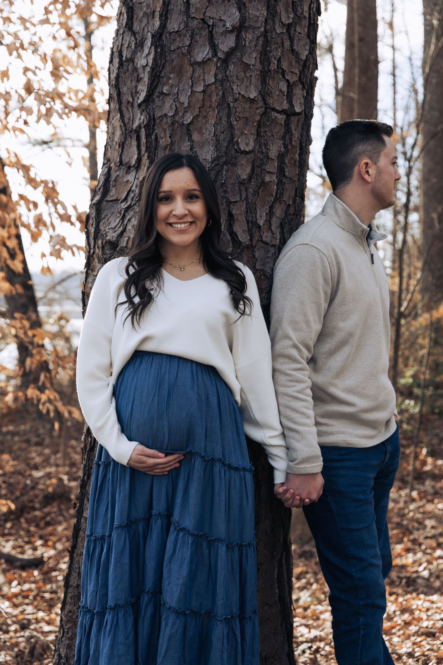 Maternity photos of couple in white and navy by large pine trees.