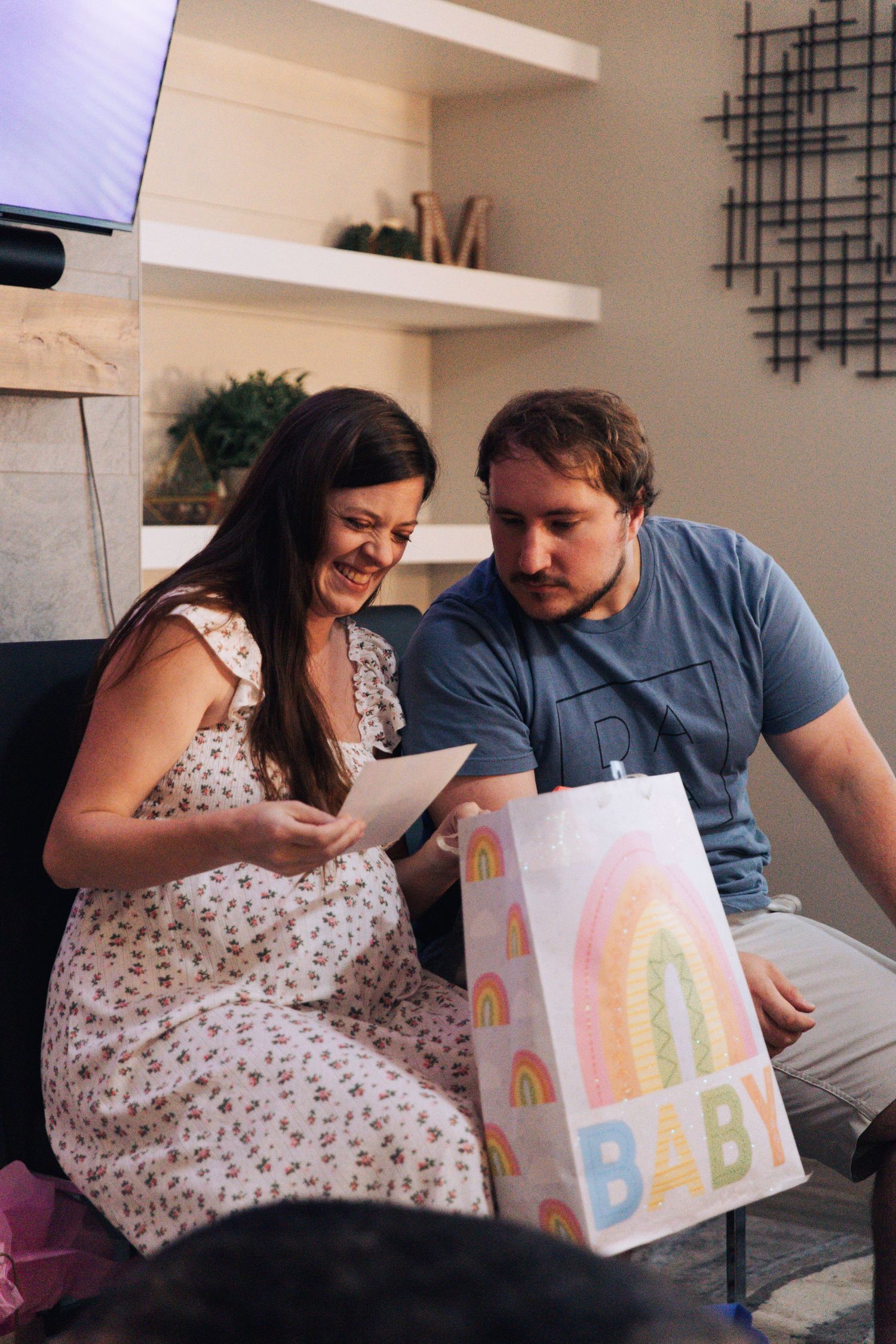 A couple sits together looking at a colorful baby-themed card in their living room.