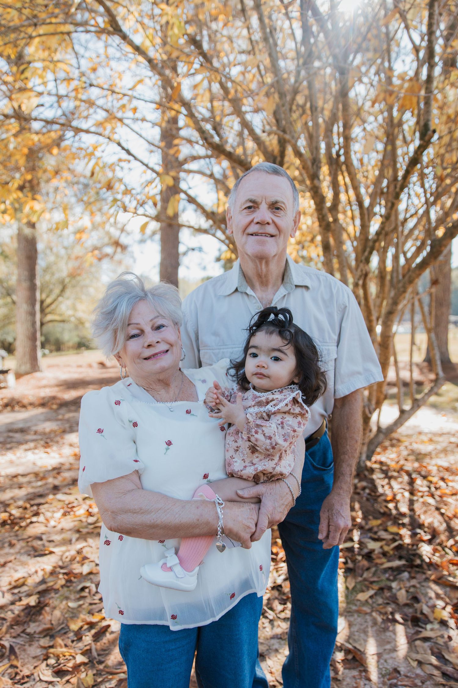 Multi-generational family poses together in a wooded autumn setting with fallen leaves.