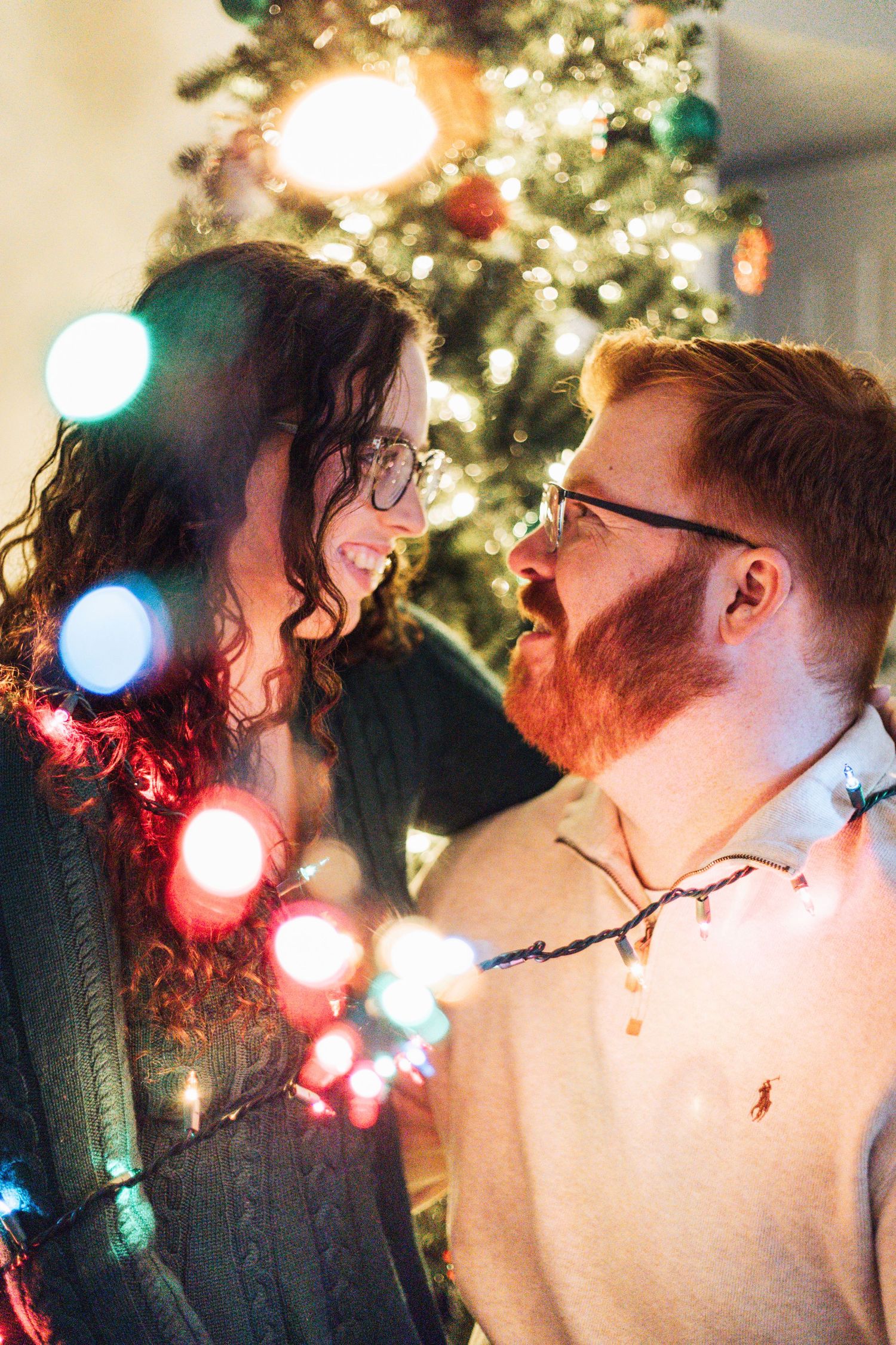 A couple shares a romantic kiss in front of a glowing Christmas tree with colorful bokeh lights in the background.