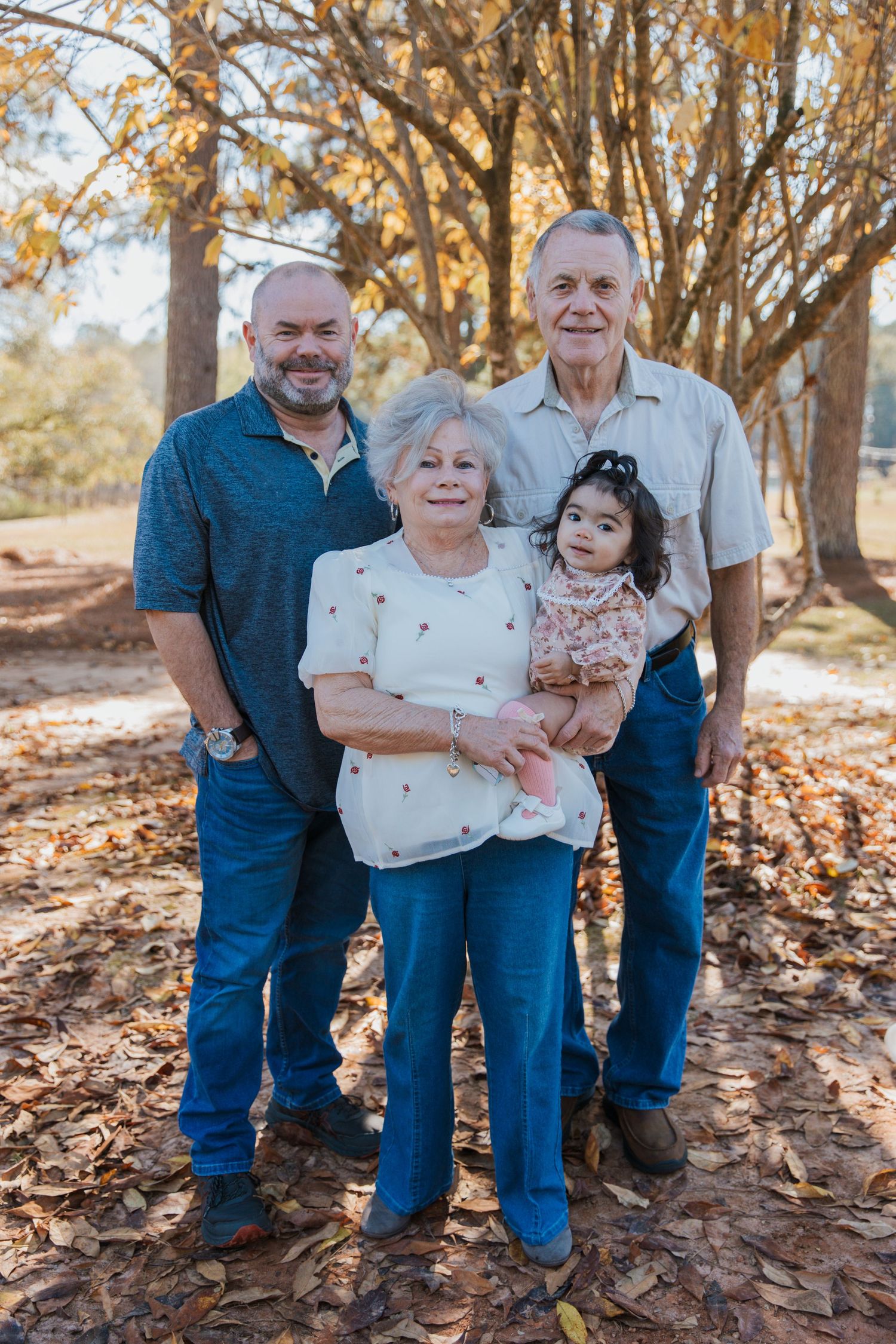 Multi-generational family gathering poses together outdoors among autumn trees and fallen leaves.