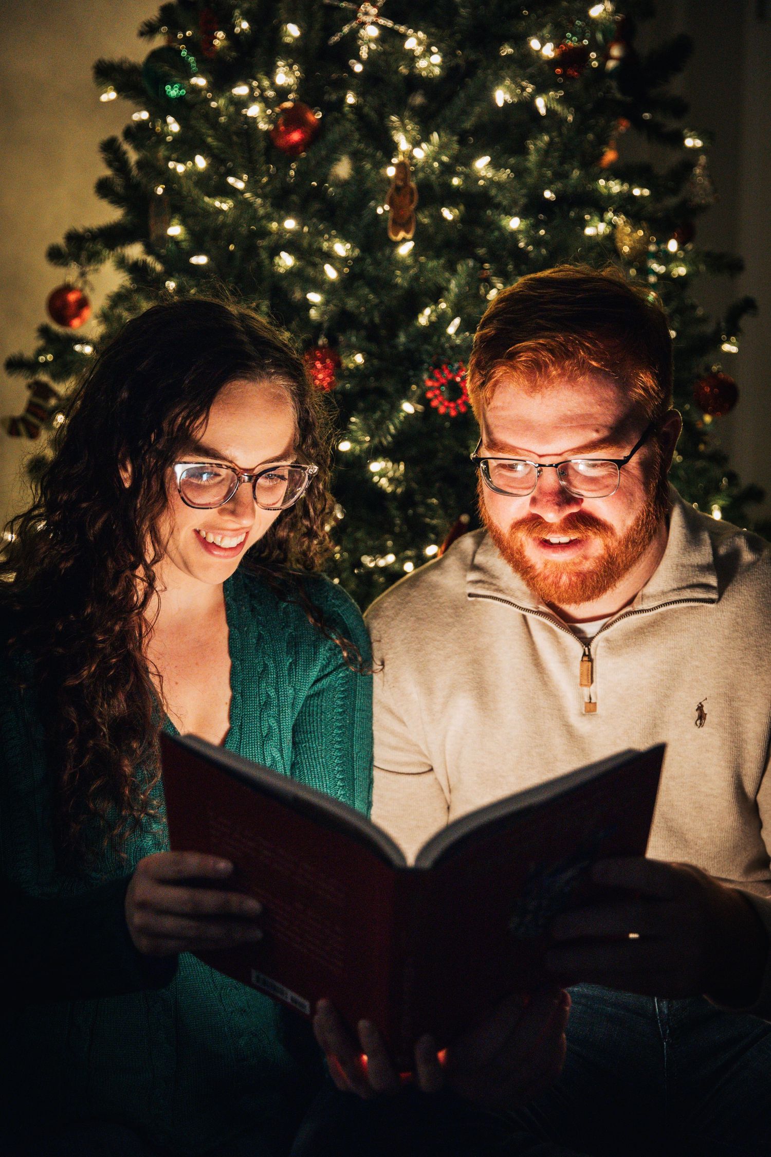 Two people reading a book together in front of a decorated Christmas tree with twinkling lights in a cozy setting.