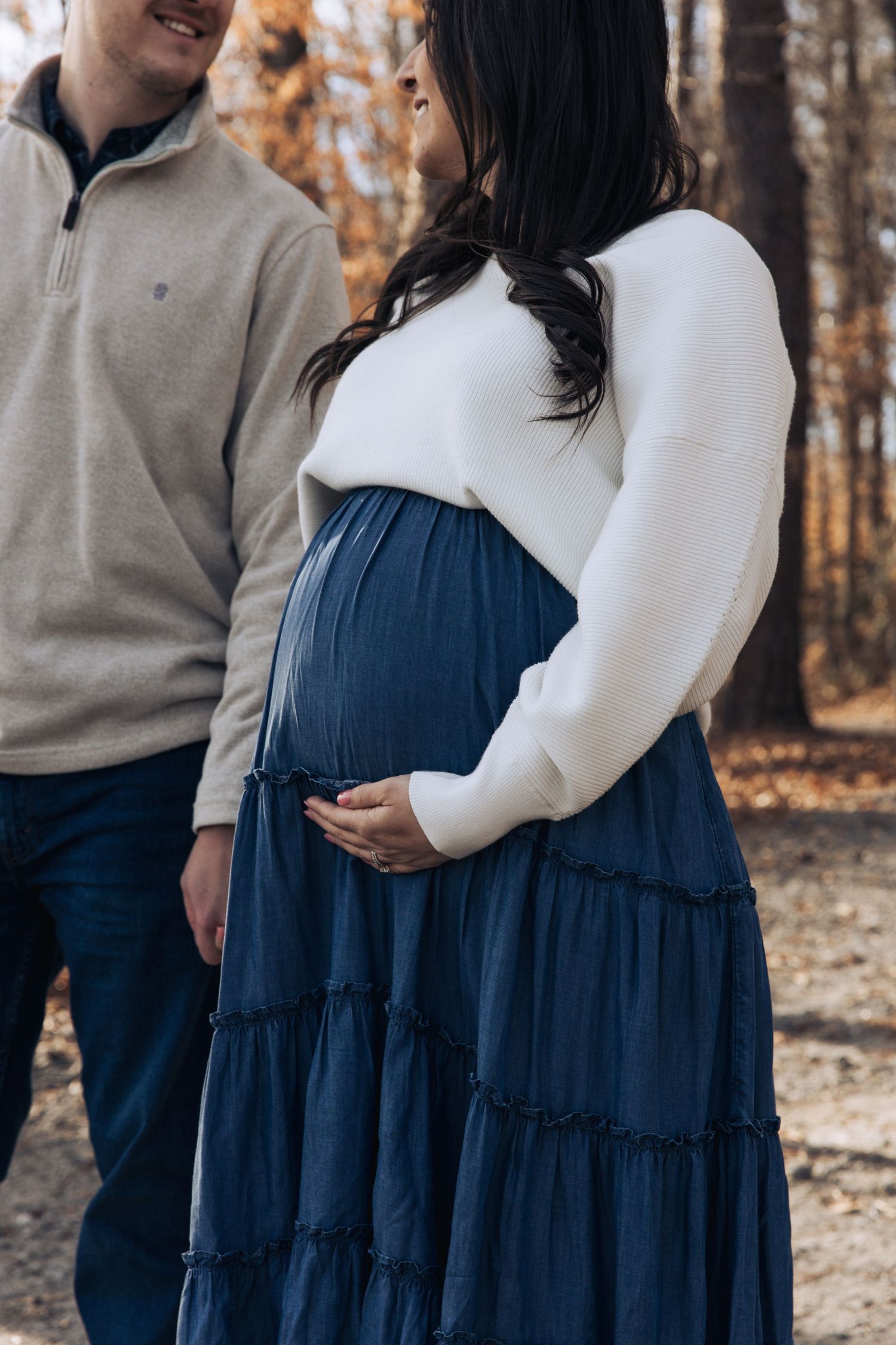 Expectant couple in autumn woods with woman wearing white sweater and navy tiered maxi dress cradling baby bump.