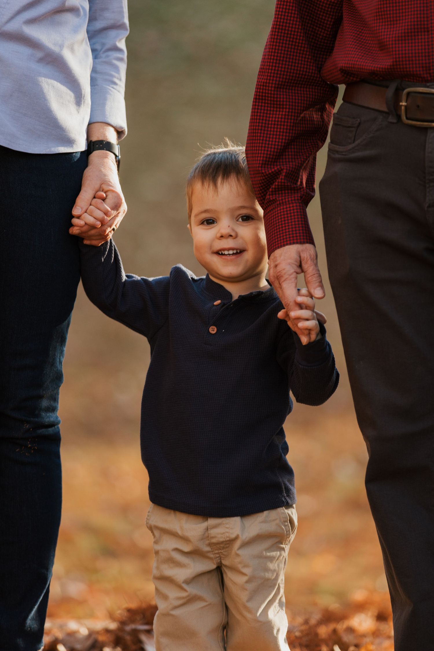 A small child in a navy sweater and khaki pants holds hands with adults while walking through autumn leaves.