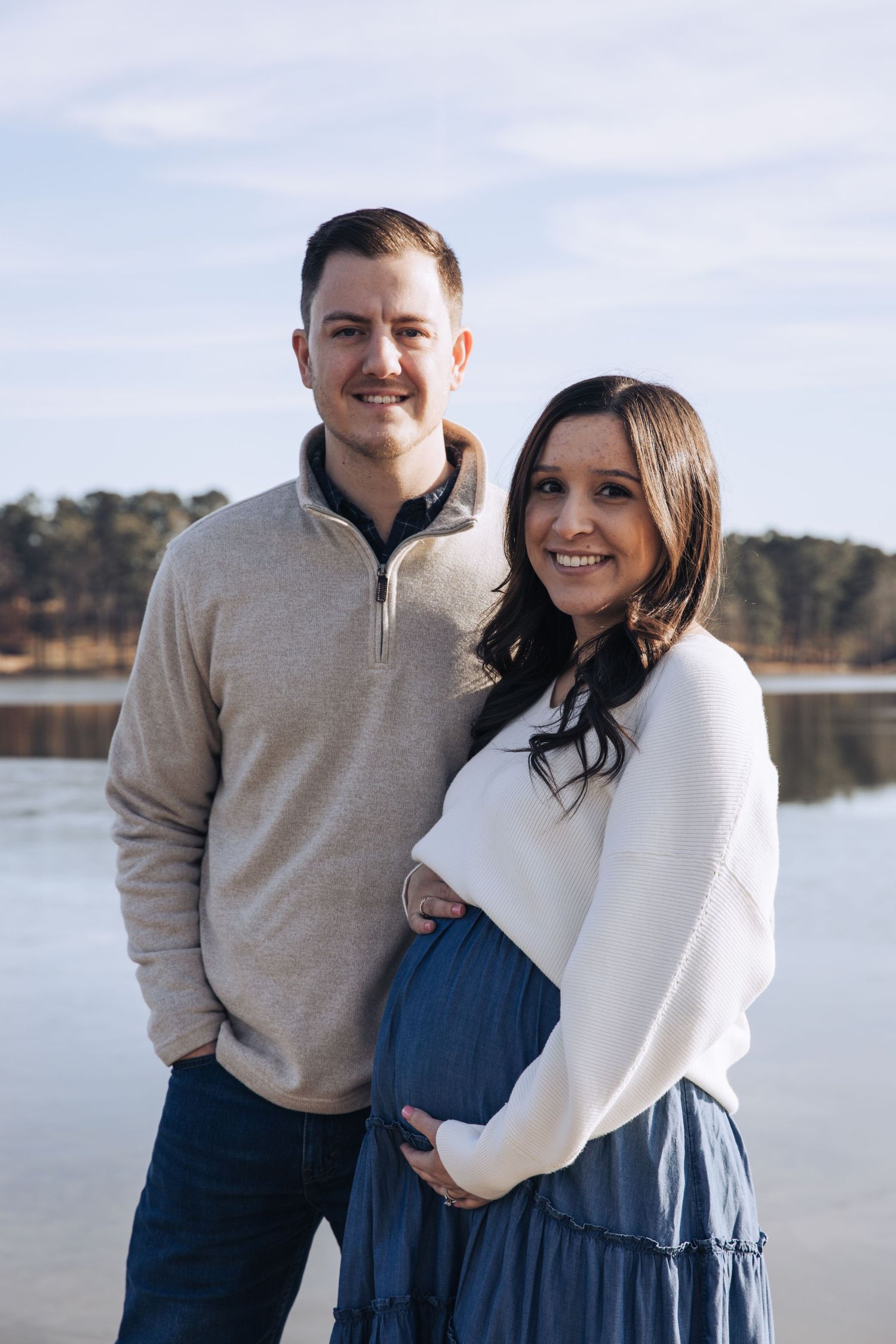 A couple poses together outdoors near a lake wearing coordinating beige and navy blue outfits in a romantic photoshoot.