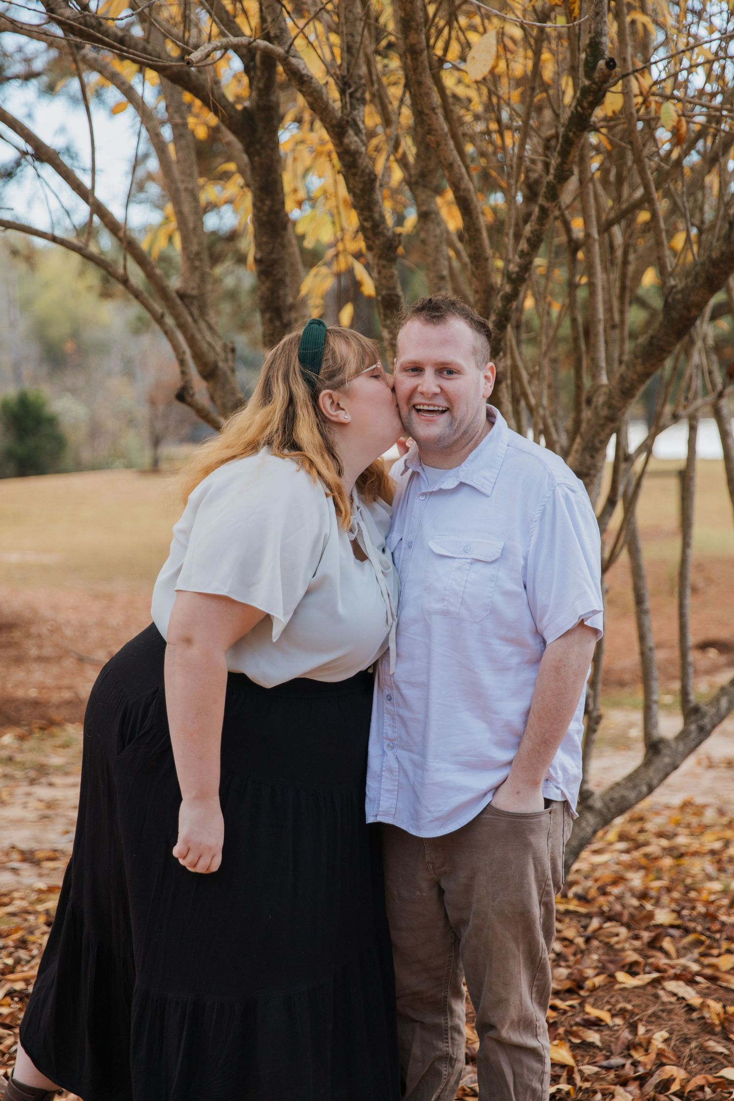 A couple shares tender moments together while standing among autumn trees with fallen leaves on the ground.