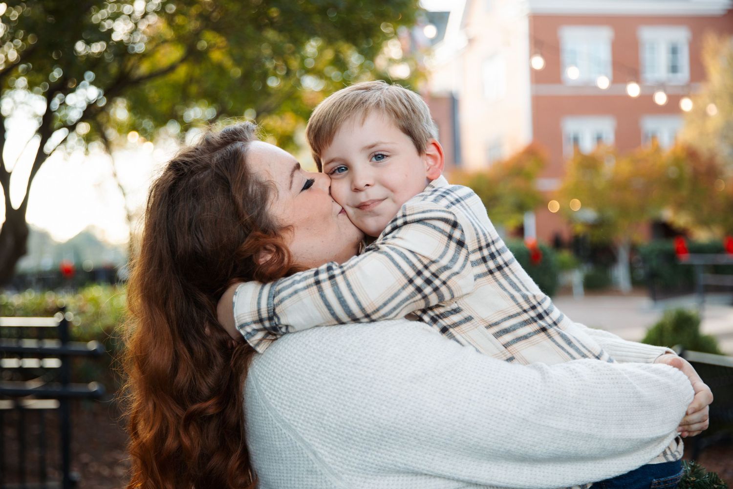 Two people embrace in a warm hug outdoors during autumn with brick buildings in the background.