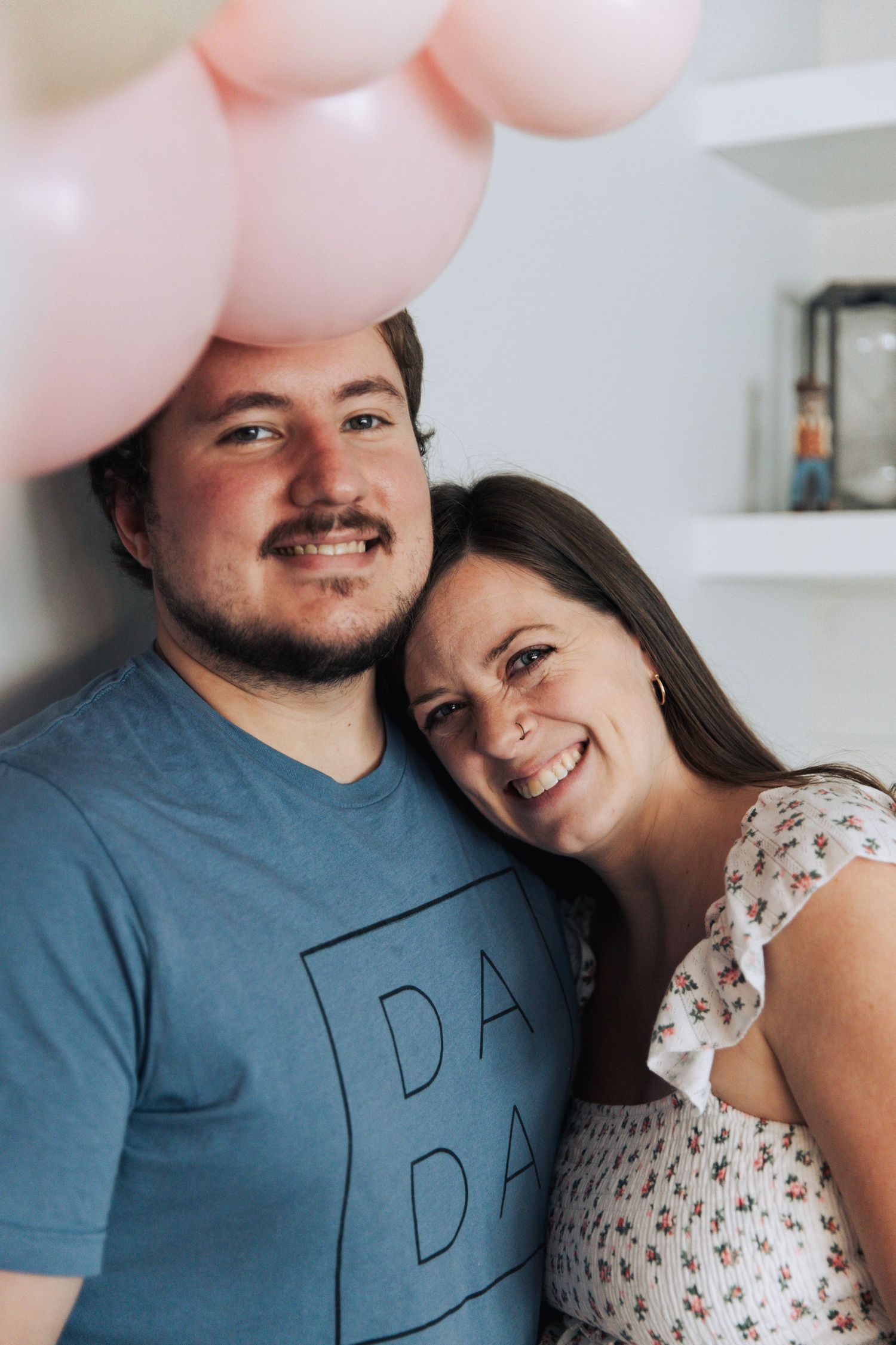 A couple smiles together in front of a white wall decorated with pink balloons.