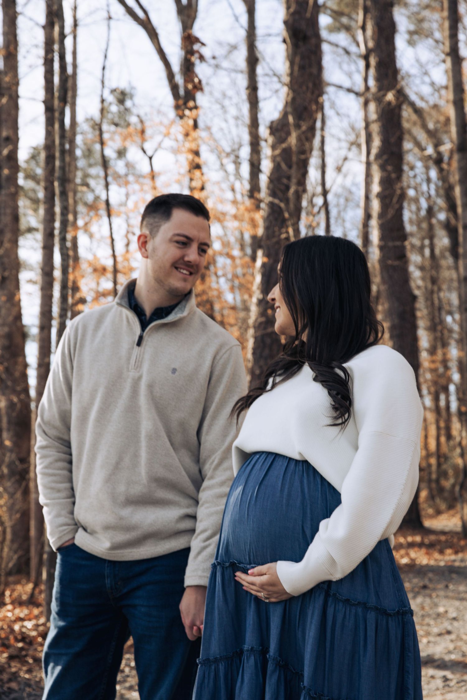 A couple in coordinating beige and navy outfits share tender moments in a wooded autumn setting.