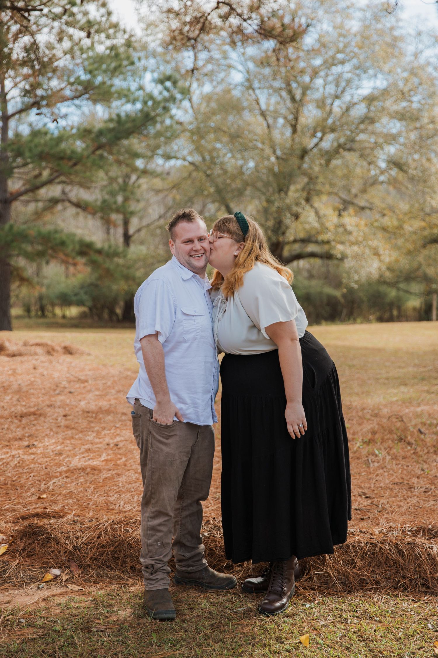 A couple shares a tender moment together in an autumn field with fallen leaves and trees in the background.