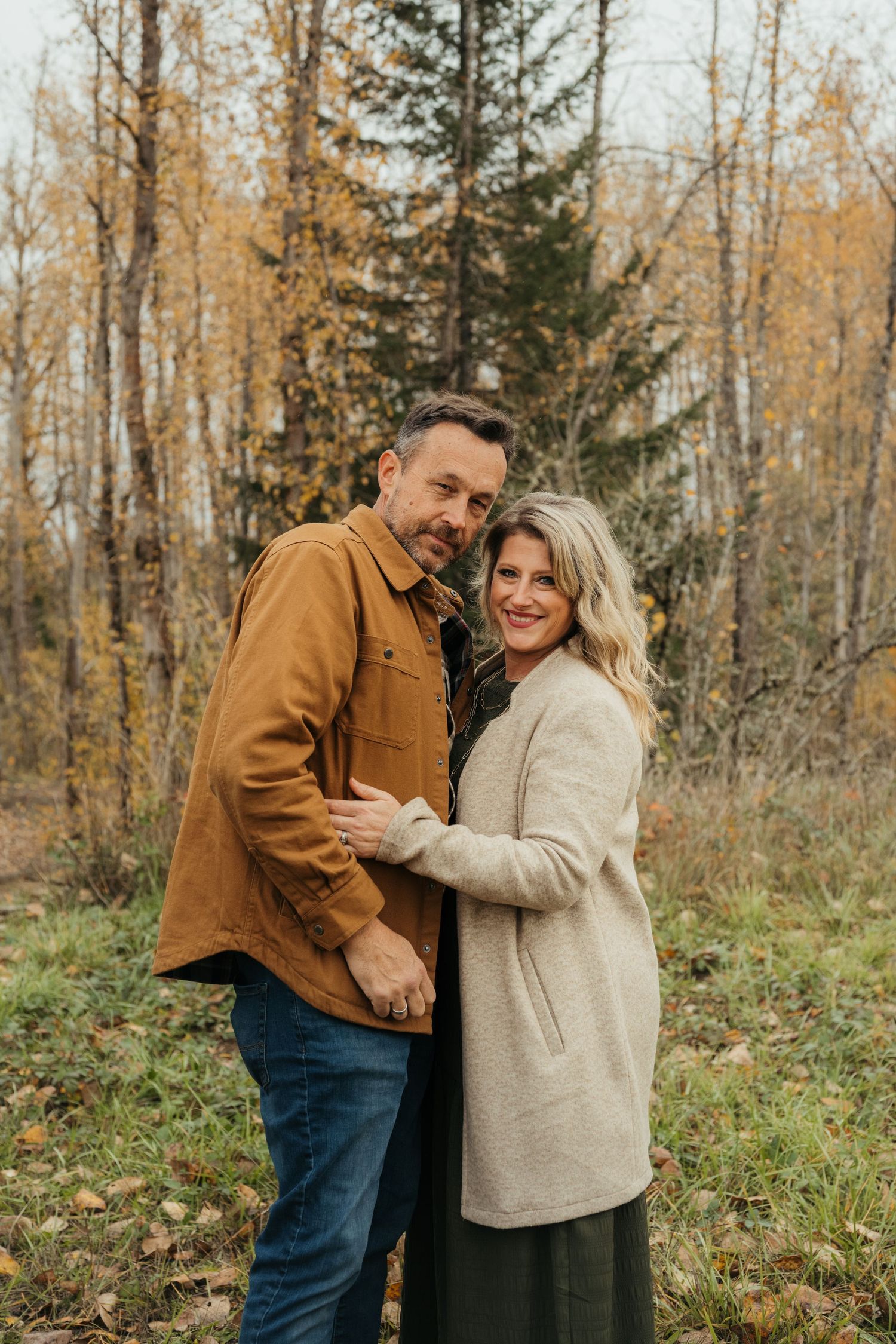 A couple in casual fall attire poses together against a backdrop of golden autumn trees.