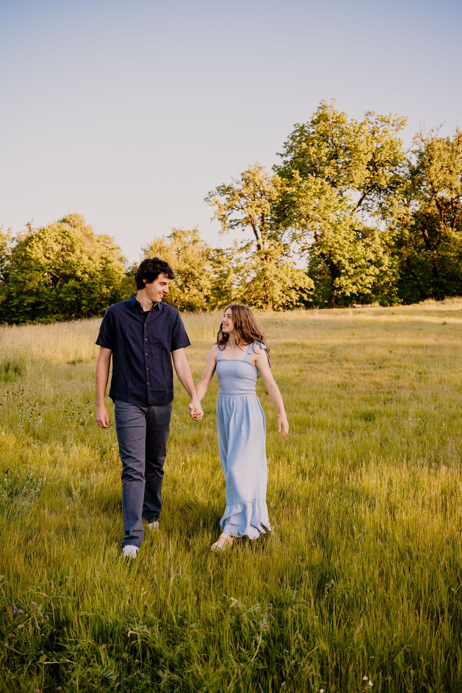 A couple holds hands walking through a sunny green field, smiling at each other, with trees in the background.
