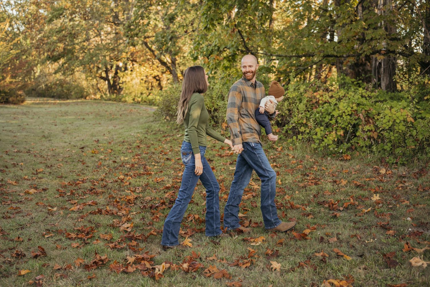 A happy family walks through an autumn park, with a man carrying a baby and holding hands with a woman among fallen leaves.
