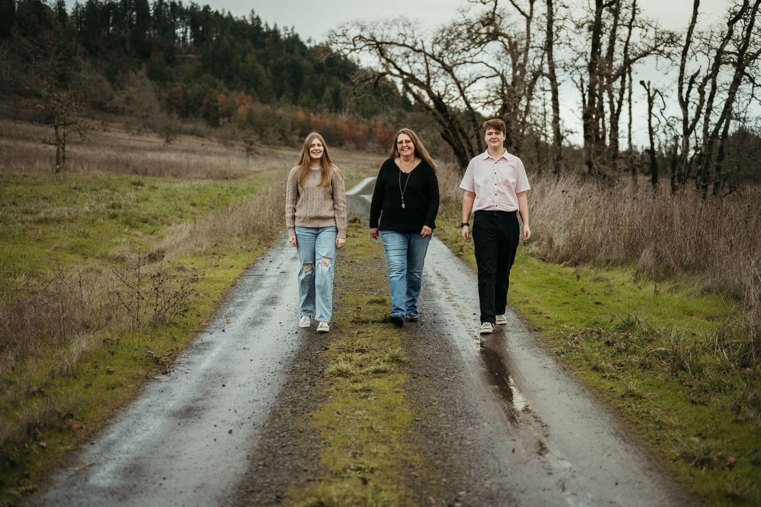 Three friends walk together down a wet gravel road surrounded by winter trees.