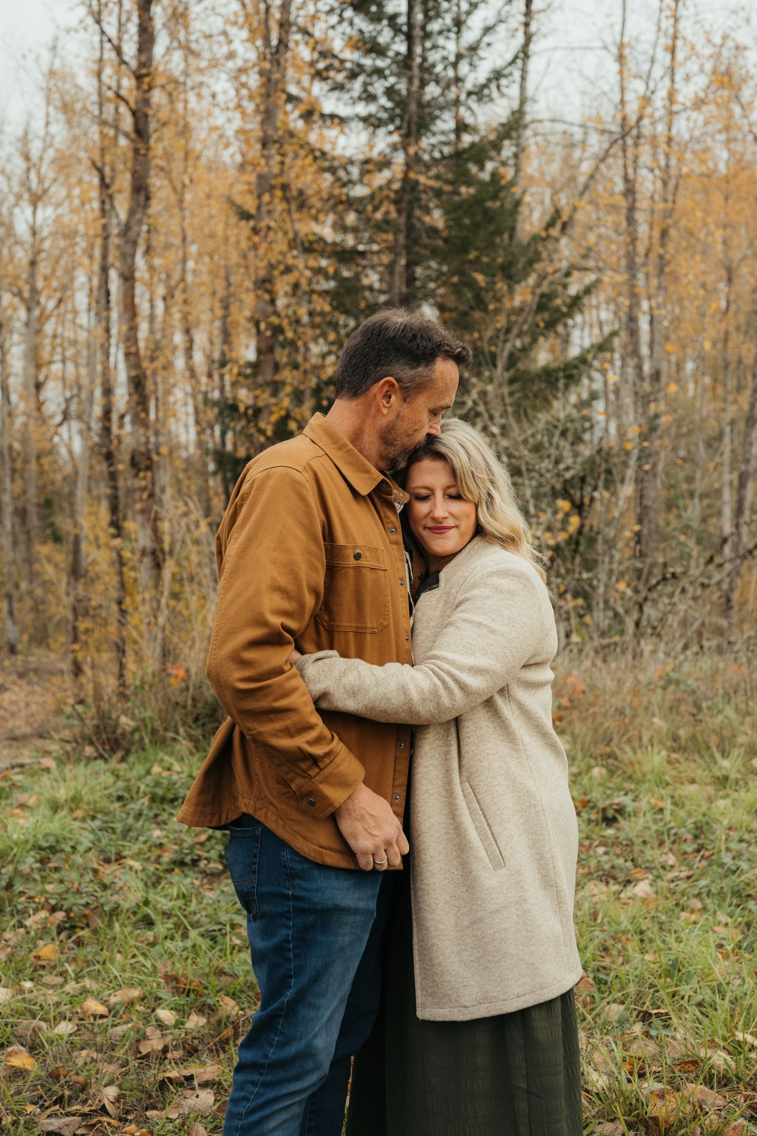 A tender embrace between a couple wearing fall clothing in a wooded setting.