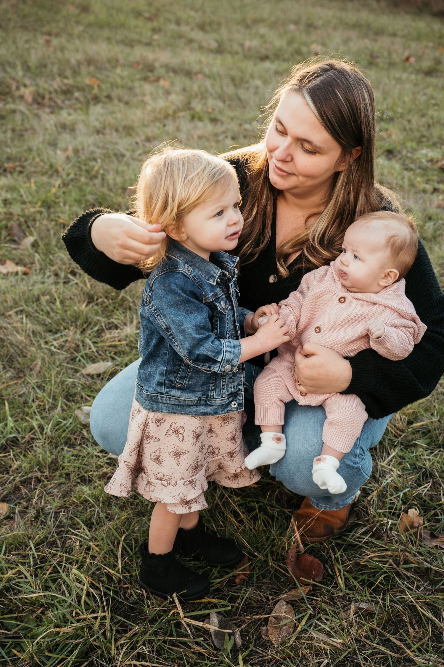 A mother crouches outdoors holding an infant in pink while touching her toddler daughter's hair in a grassy field.