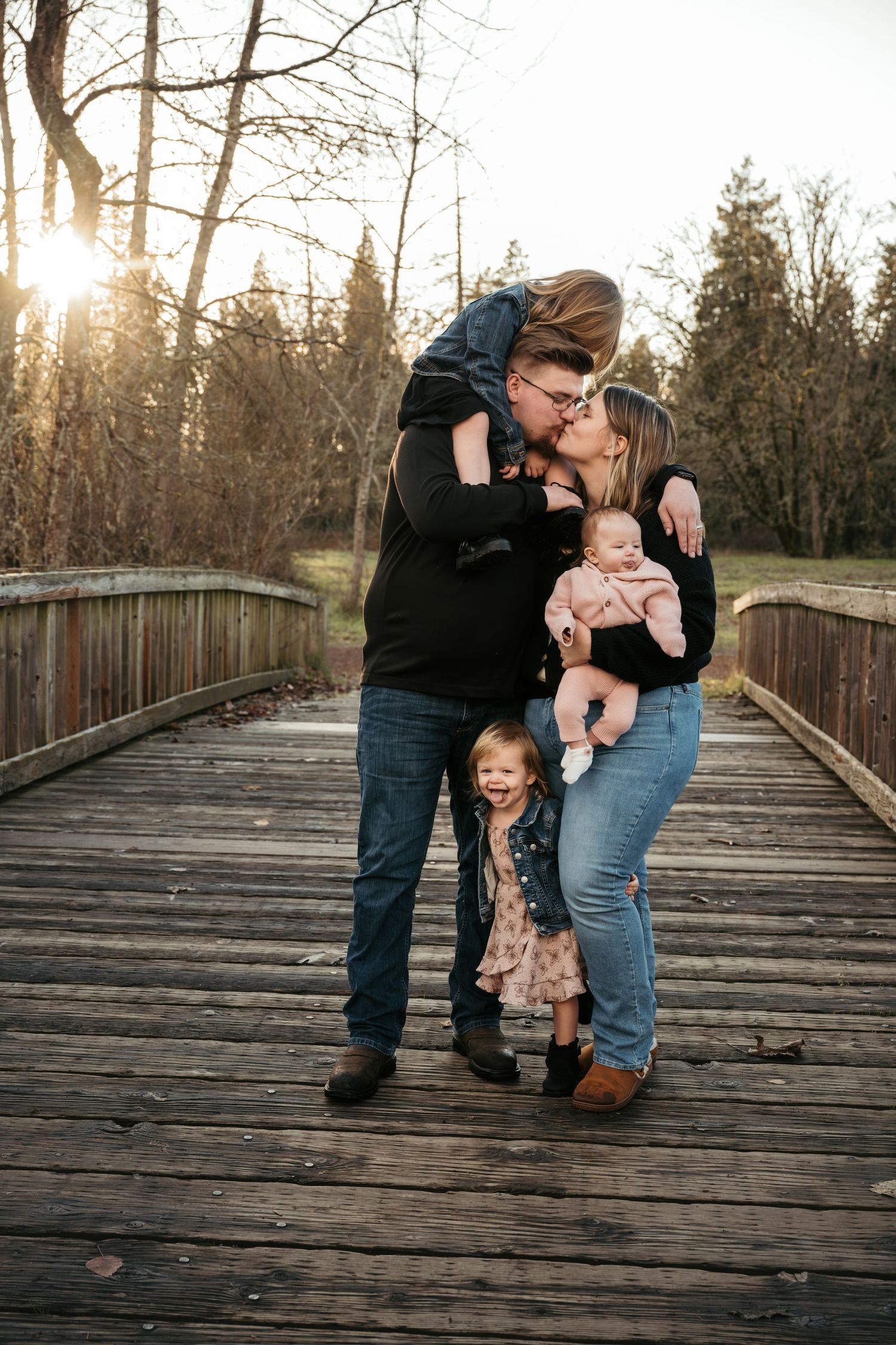 Family of four on wooden bridge at sunset, parents kissing while holding baby and toddler daughter smiling below.