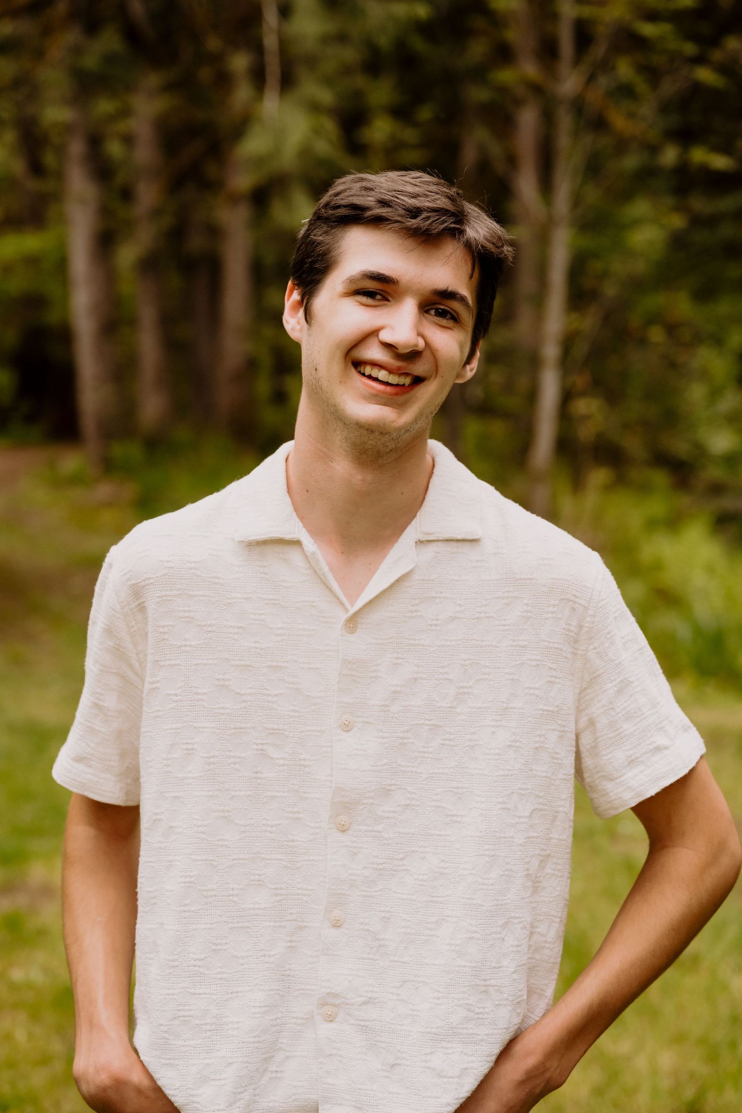 Smiling young man in a white button-up shirt standing outdoors in a lush green forest setting.