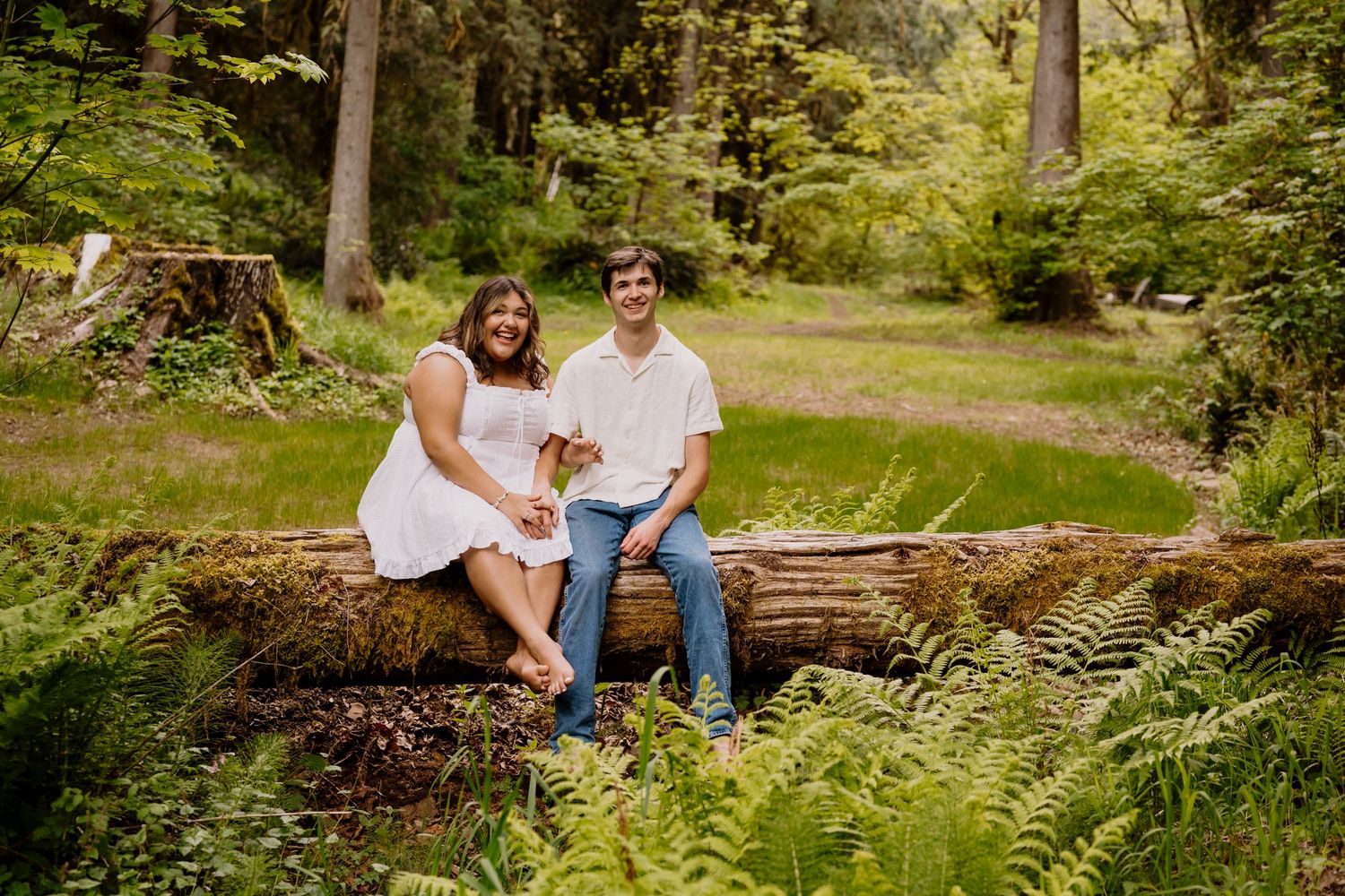Couple sitting on a fallen log in a lush green forest, woman in white dress, man in white shirt and jeans.