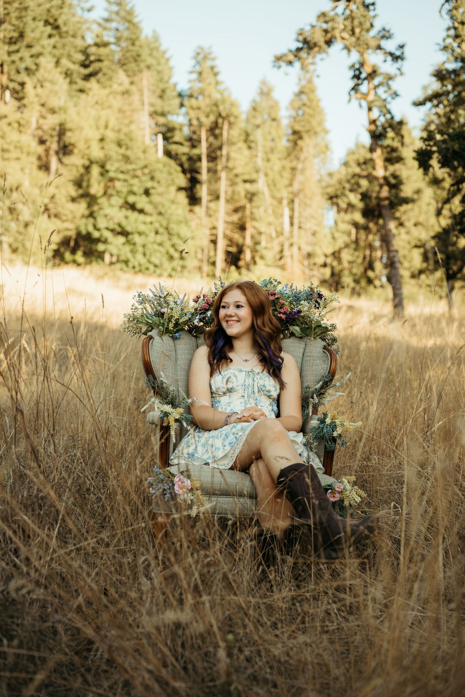 Vintage armchair with floral decorations sits in golden field with tall pine trees in background at sunset.