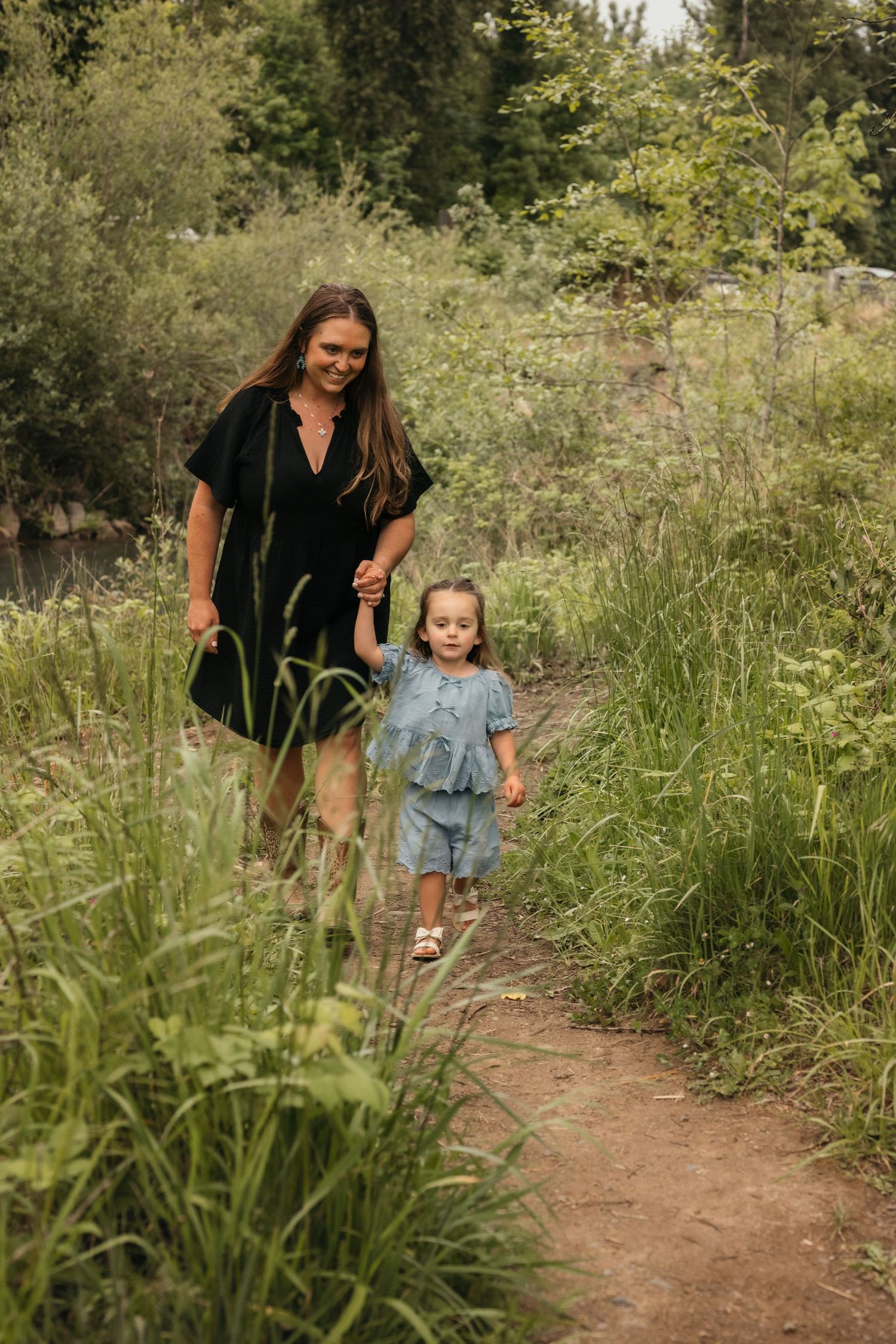 Woman in black dress holds hands with young girl in blue outfit while walking on a nature trail surrounded by greenery.