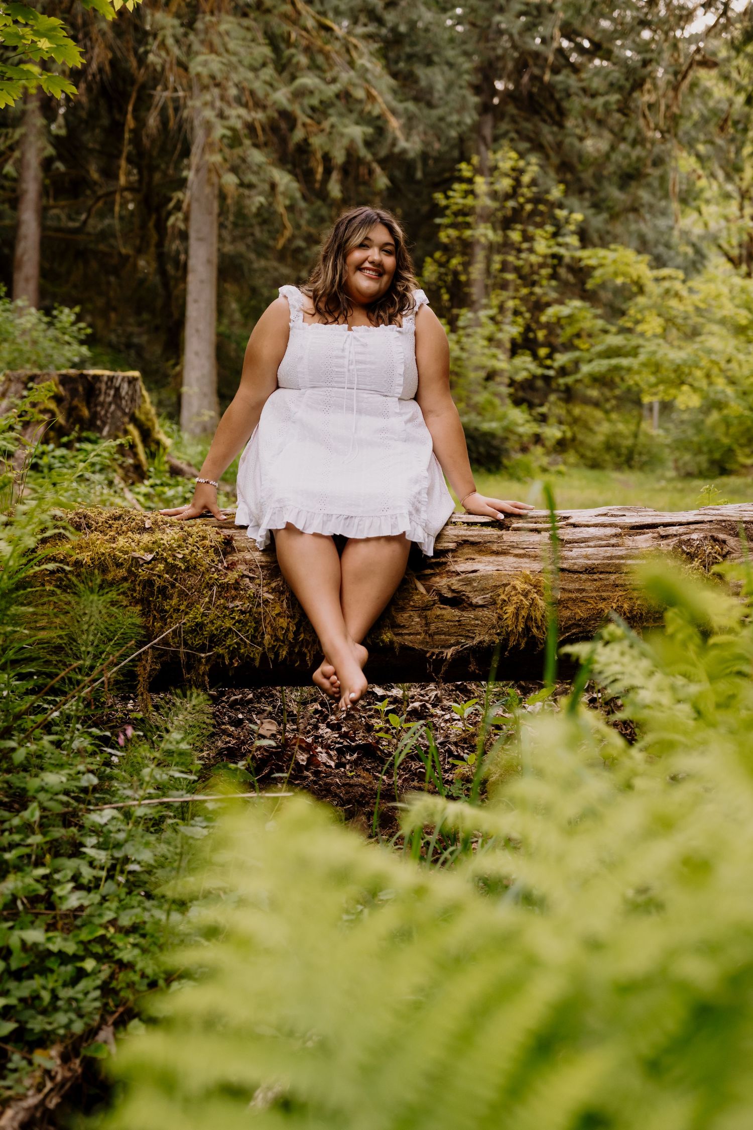 Woman in white dress sitting barefoot on mossy log in lush green forest setting.