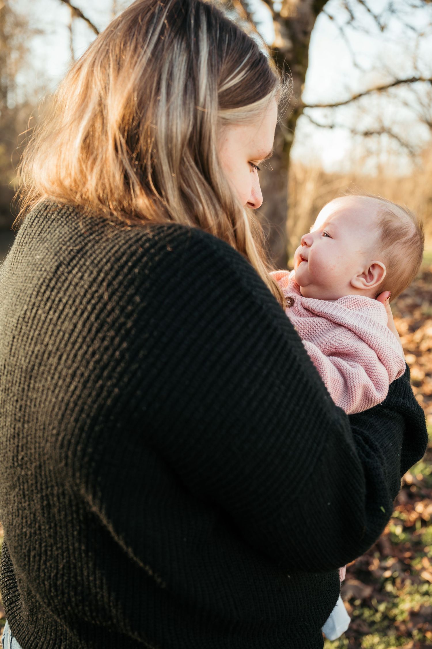A mother in a black sweater tenderly holds her newborn baby dressed in pink outdoors in warm sunlight.