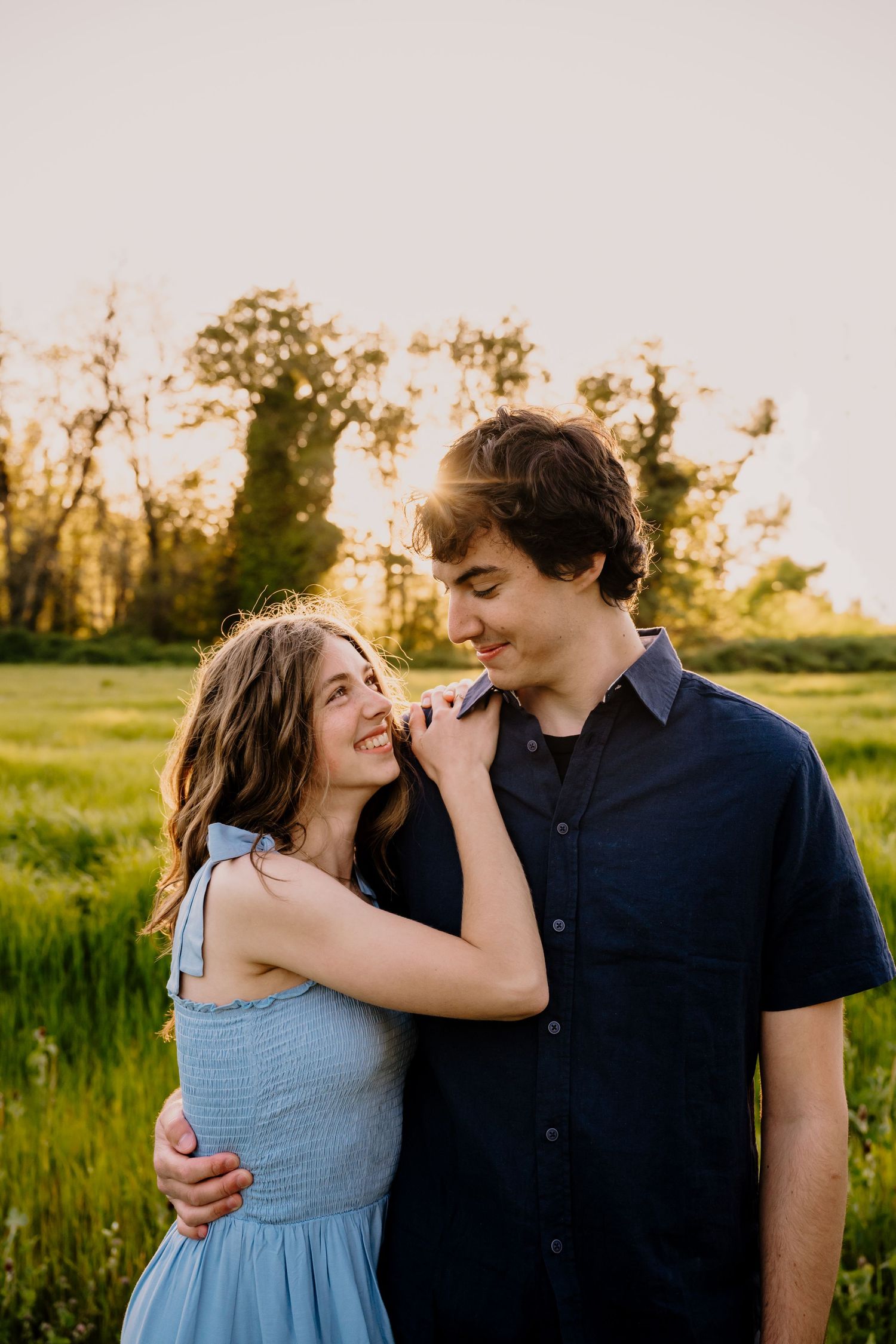 A young couple embraces in a sunlit green field at golden hour, the woman in a blue dress smiling warmly.
