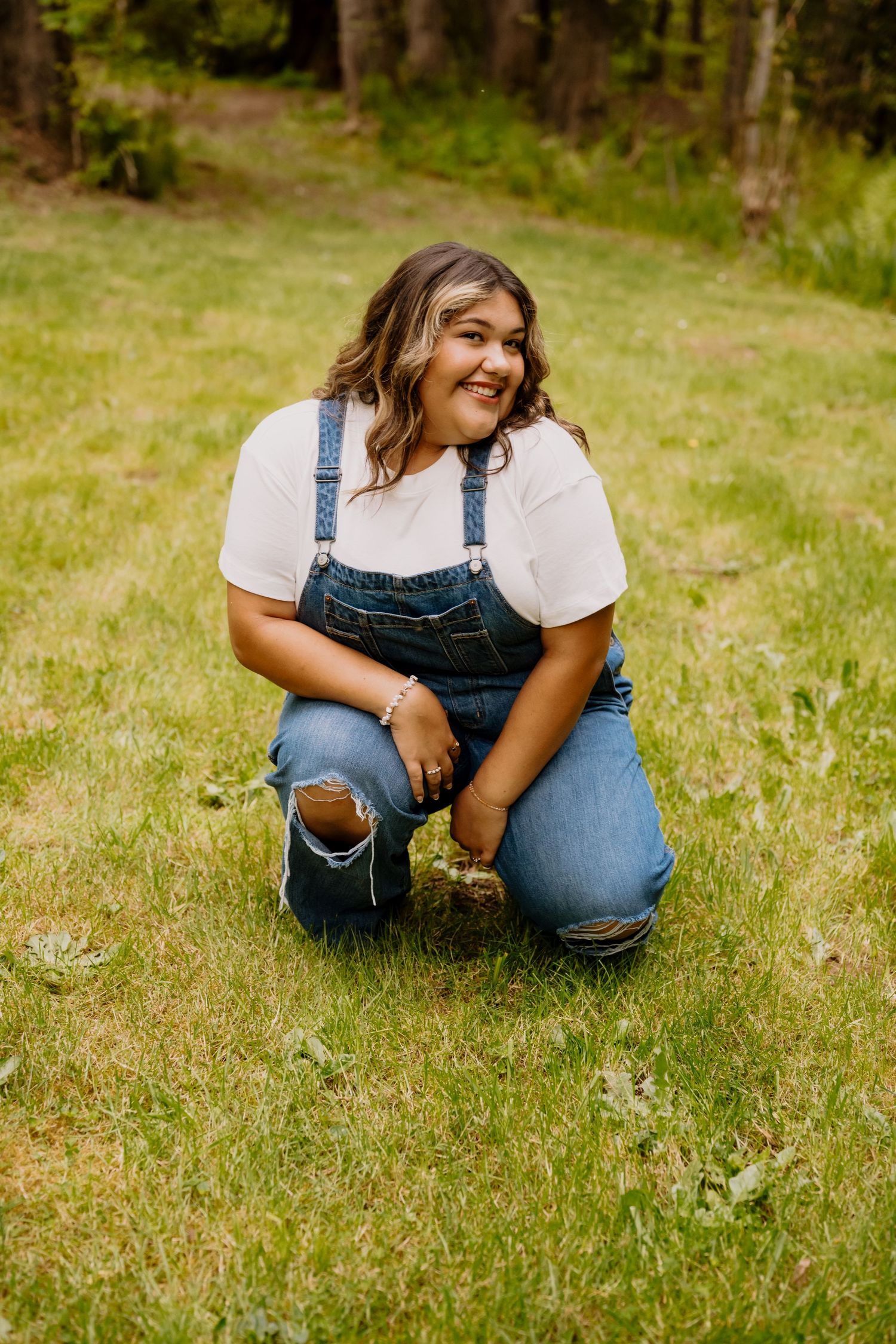 Young woman in denim overalls and white tee smiling while crouching on green grass outdoors.
