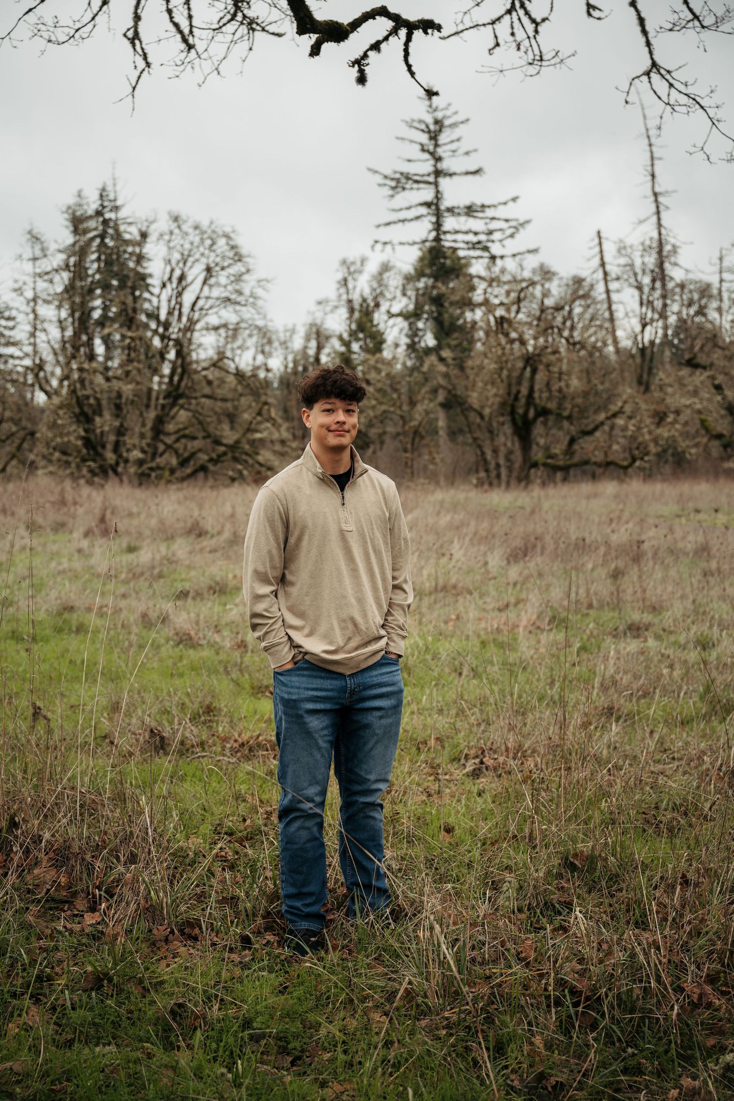 Person wearing beige sweater and jeans stands in a grassy field with bare trees in the background.