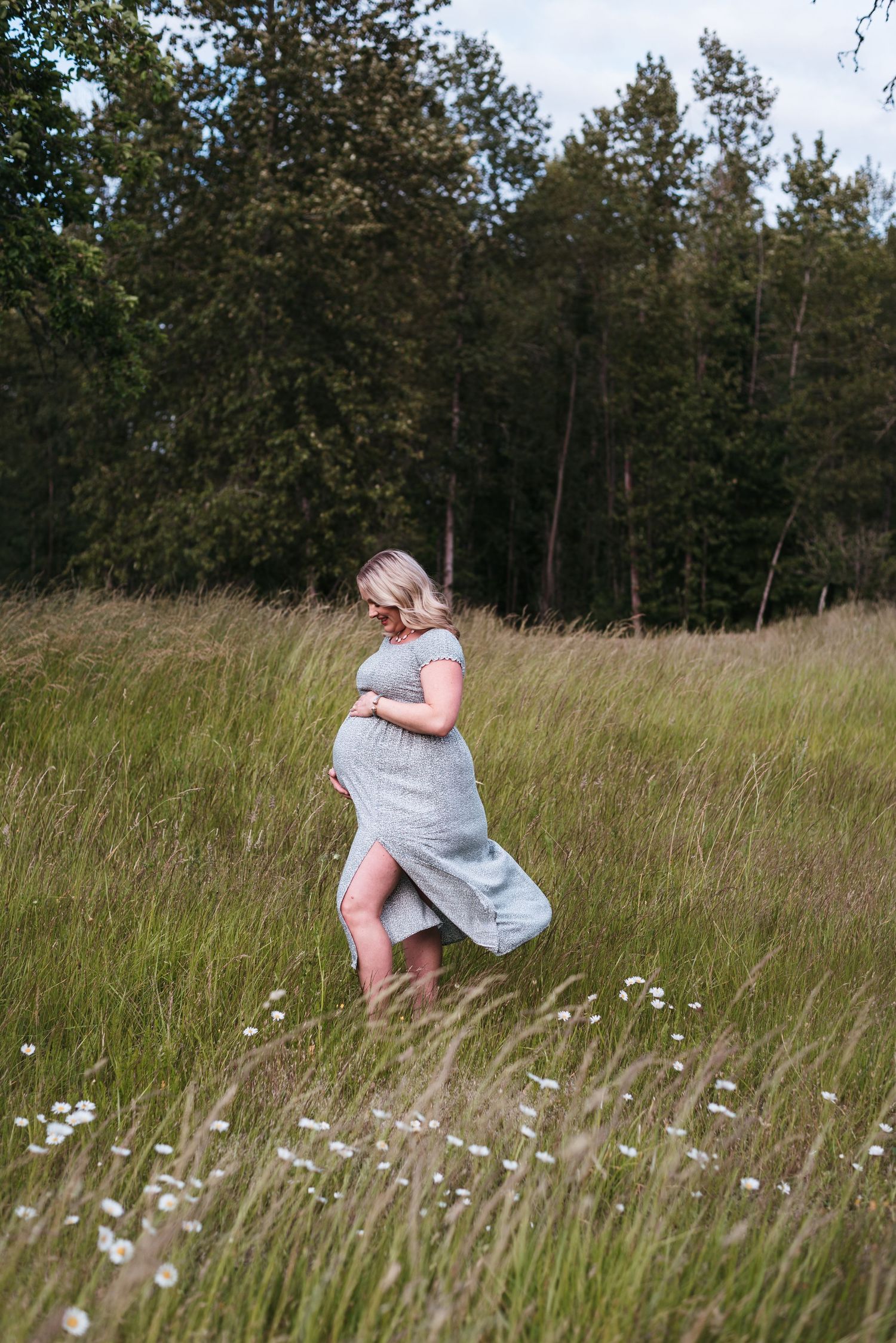 Pregnant woman in a flowing dress twirls through tall grass in a meadow.