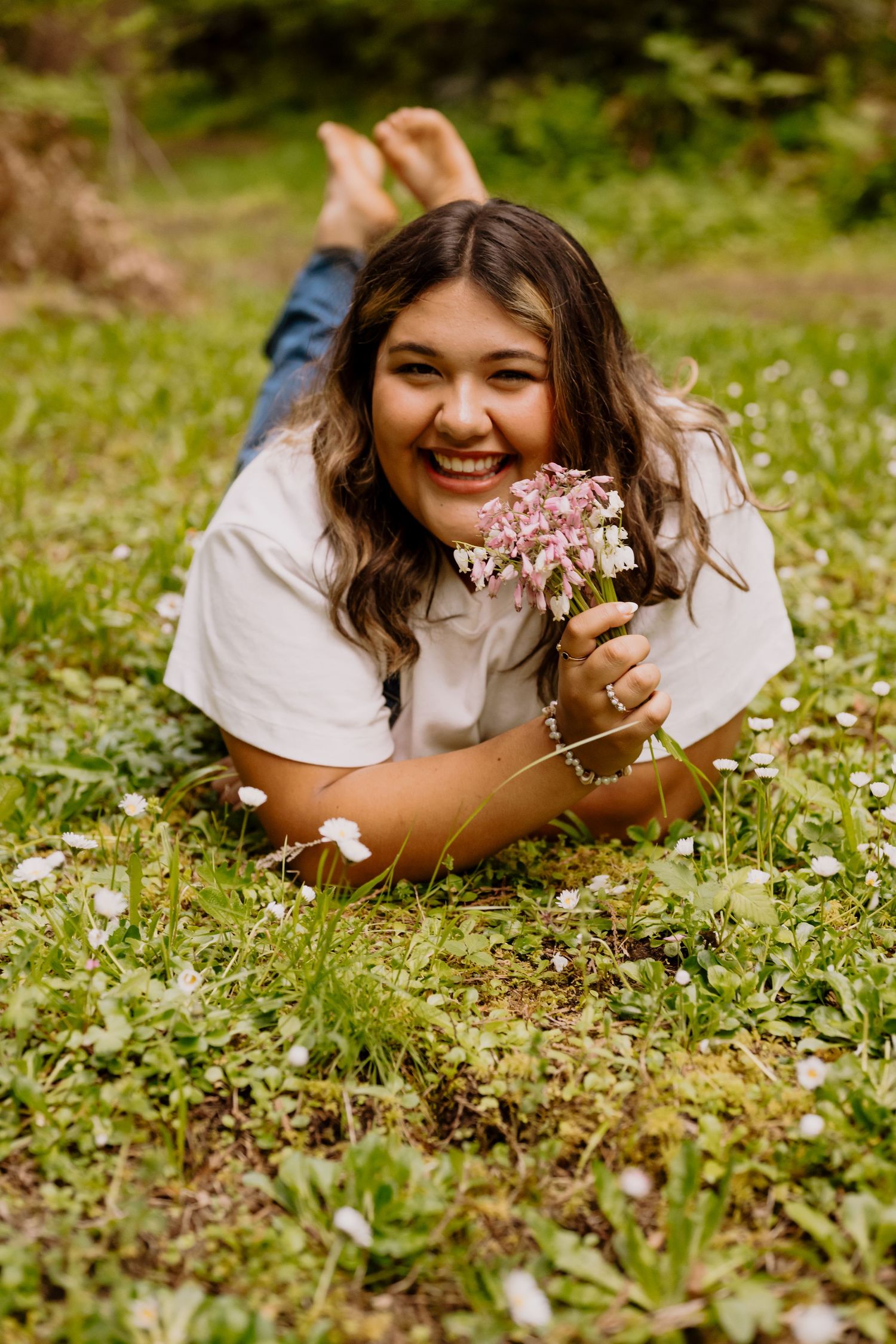 Smiling young woman lying on grass holding small pink wildflowers in a lush green outdoor meadow setting.