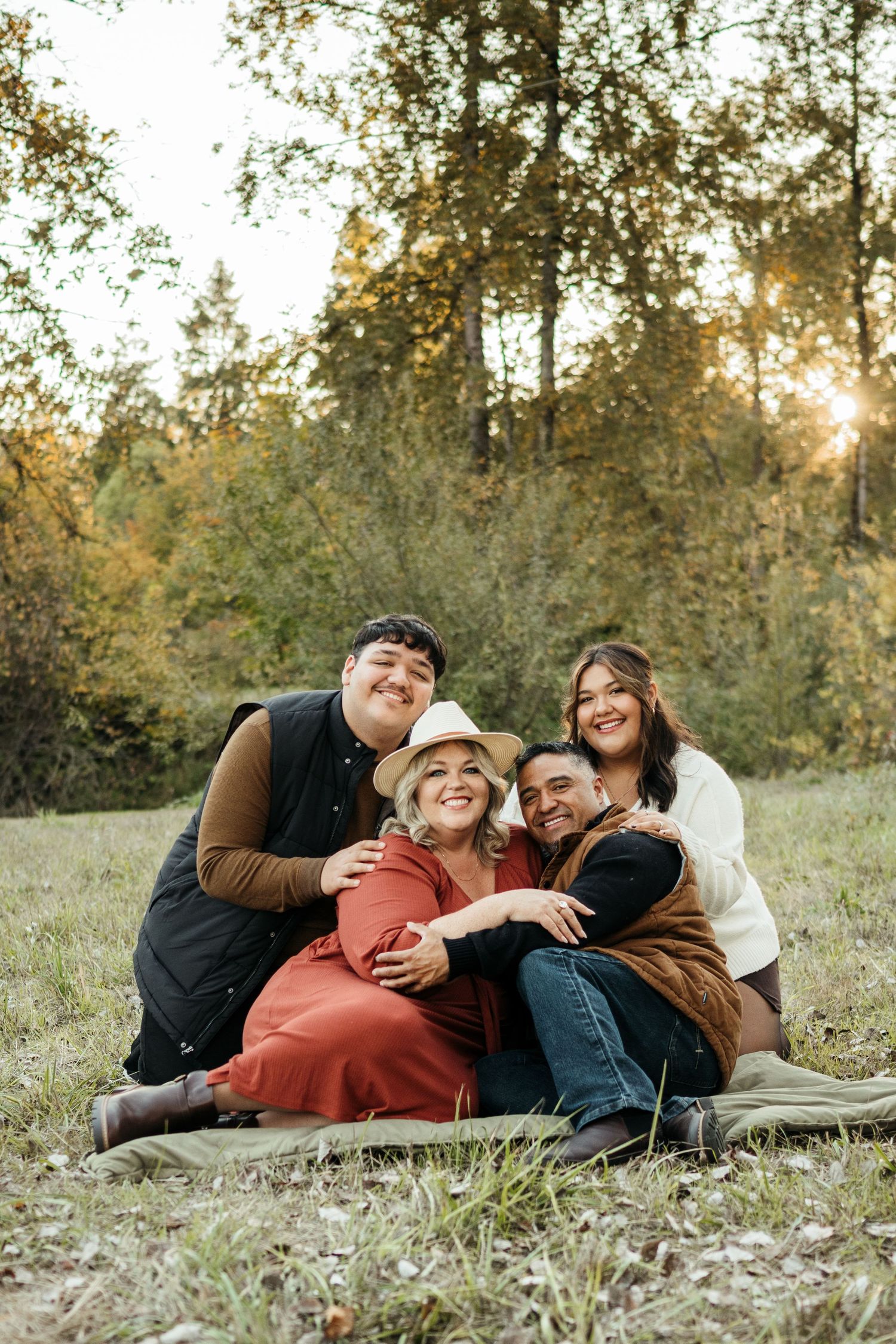 Four people sitting together on a blanket in a grassy field, smiling and hugging during a fall outdoor photo session.