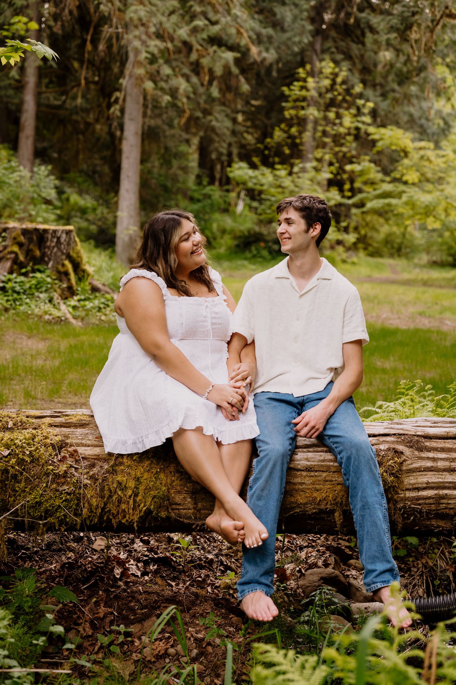 Couple sitting on a mossy log in a lush forest, smiling at each other, woman in white dress, man in jeans.