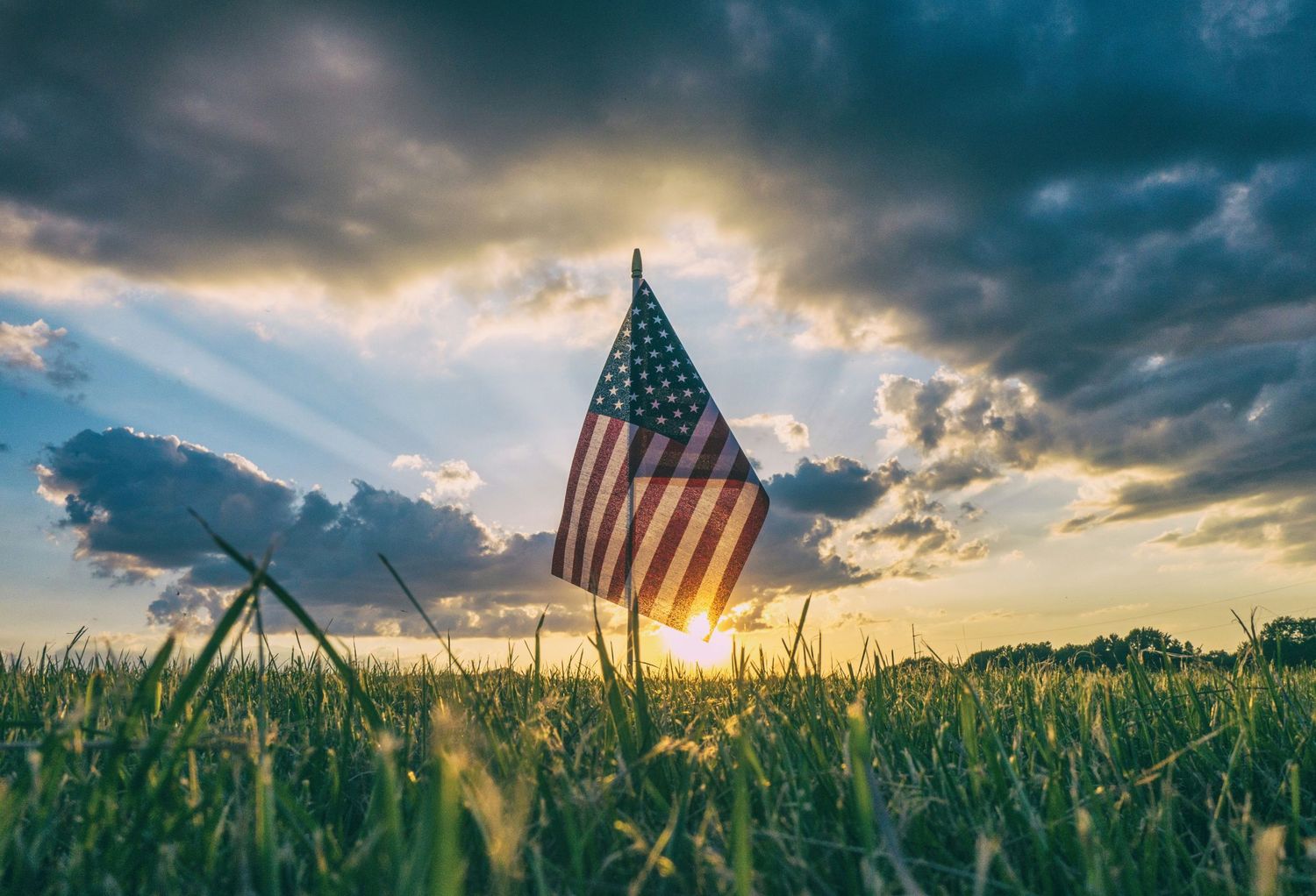 American flag standing in green grass field at sunset with dramatic cloudy sky and sunbeams shining through.