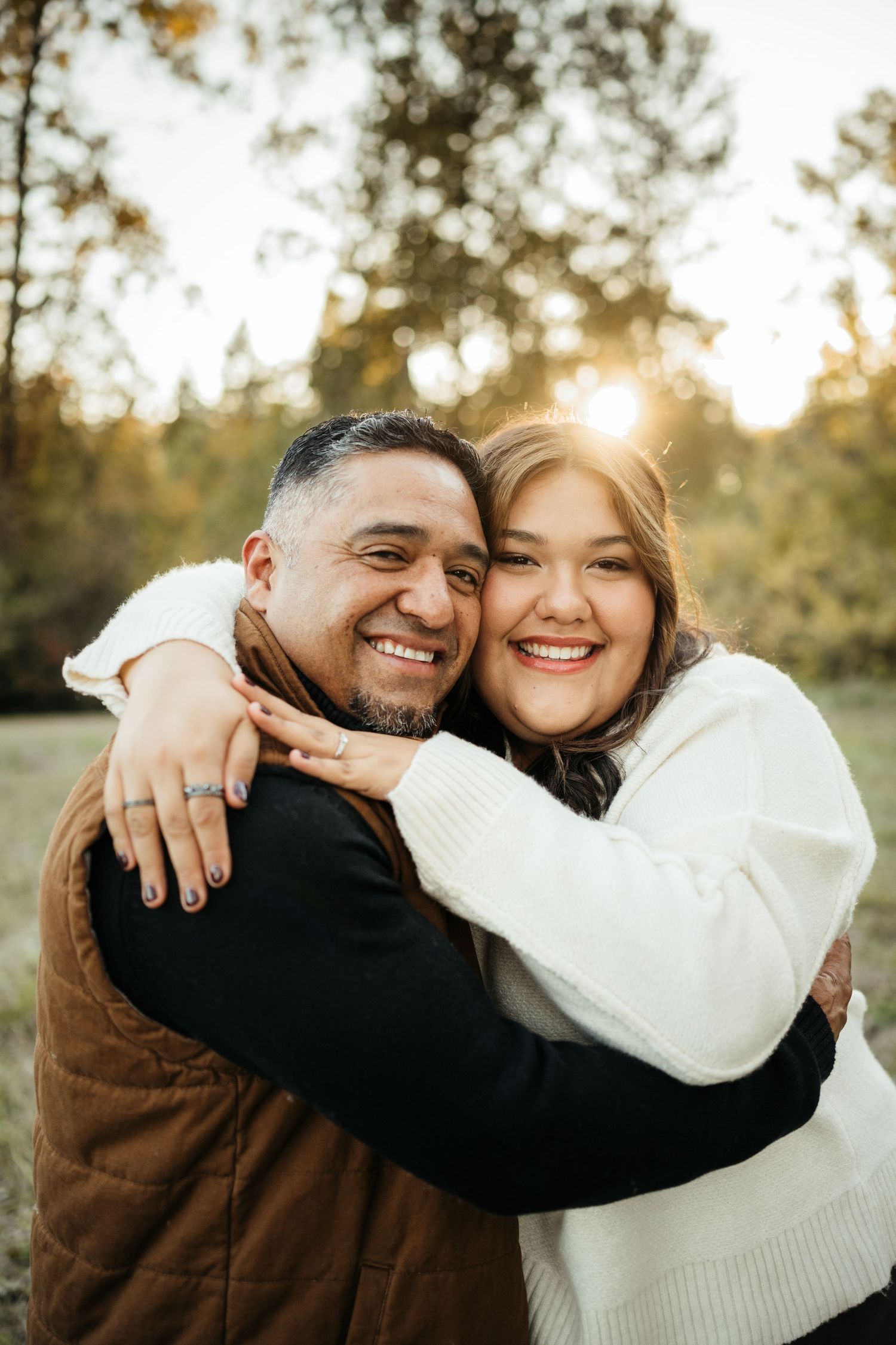 A smiling couple embraces outdoors at golden hour, with trees and sunlight in the background.