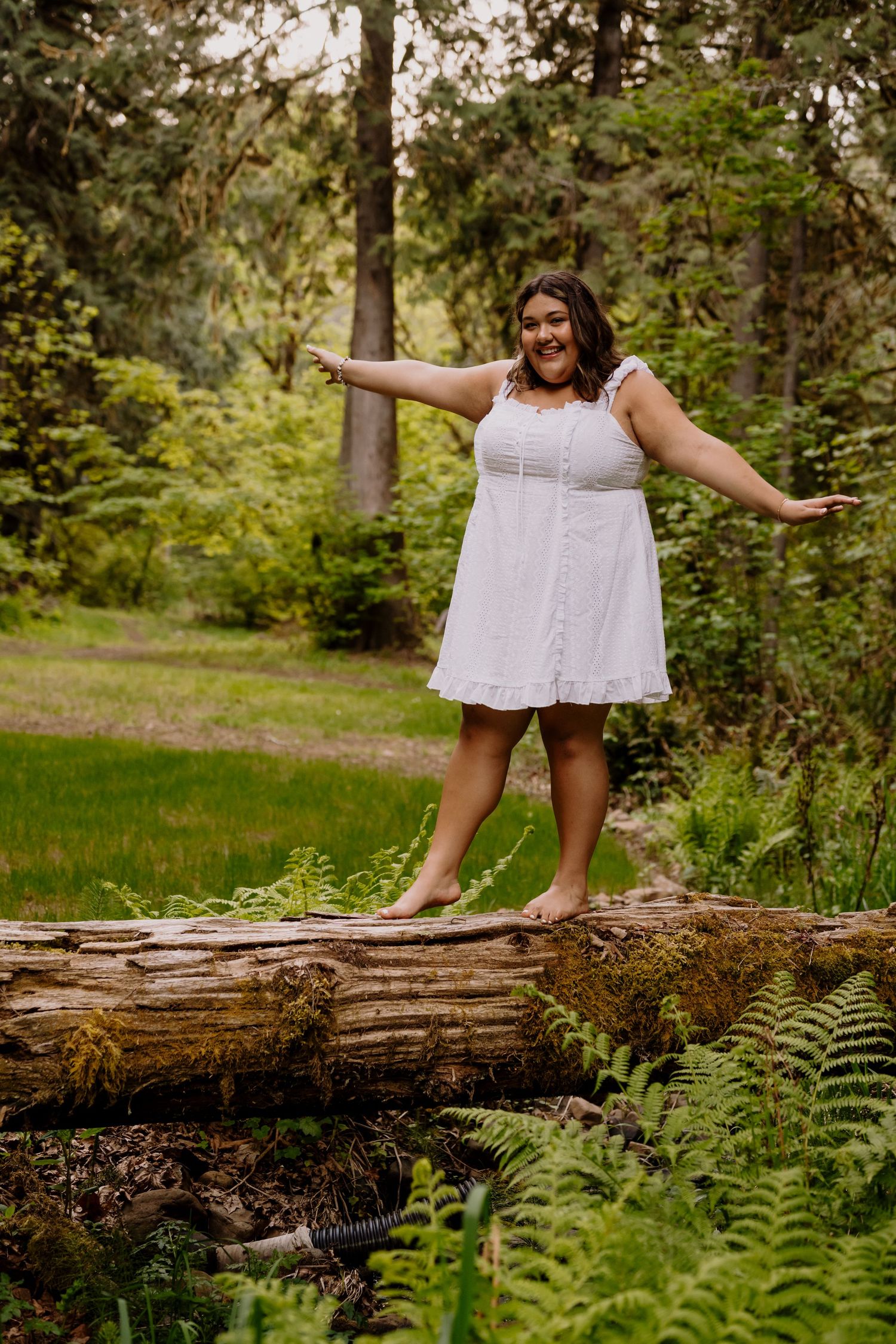 Woman in white dress balancing barefoot on fallen log in lush green forest.