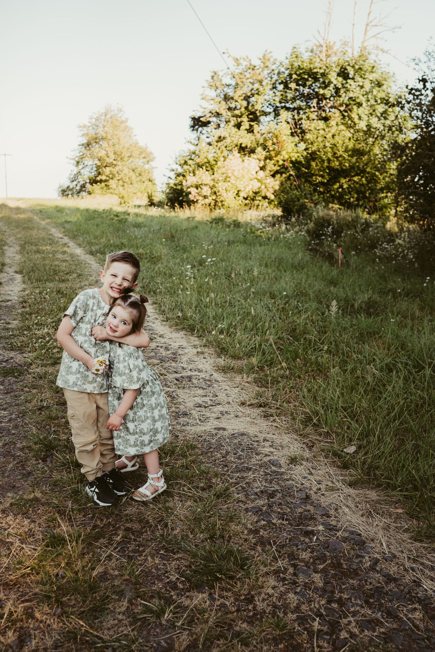 Siblings in matching floral outfits embracing on sunlit dirt path.