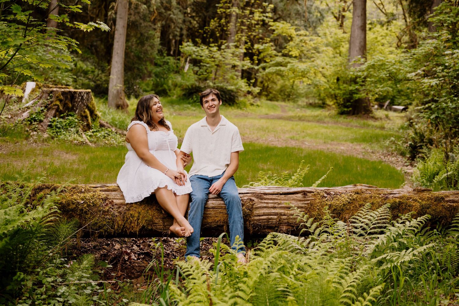 Couple sitting on mossy log in lush green forest, woman in white dress and man in white shirt and jeans, smiling.