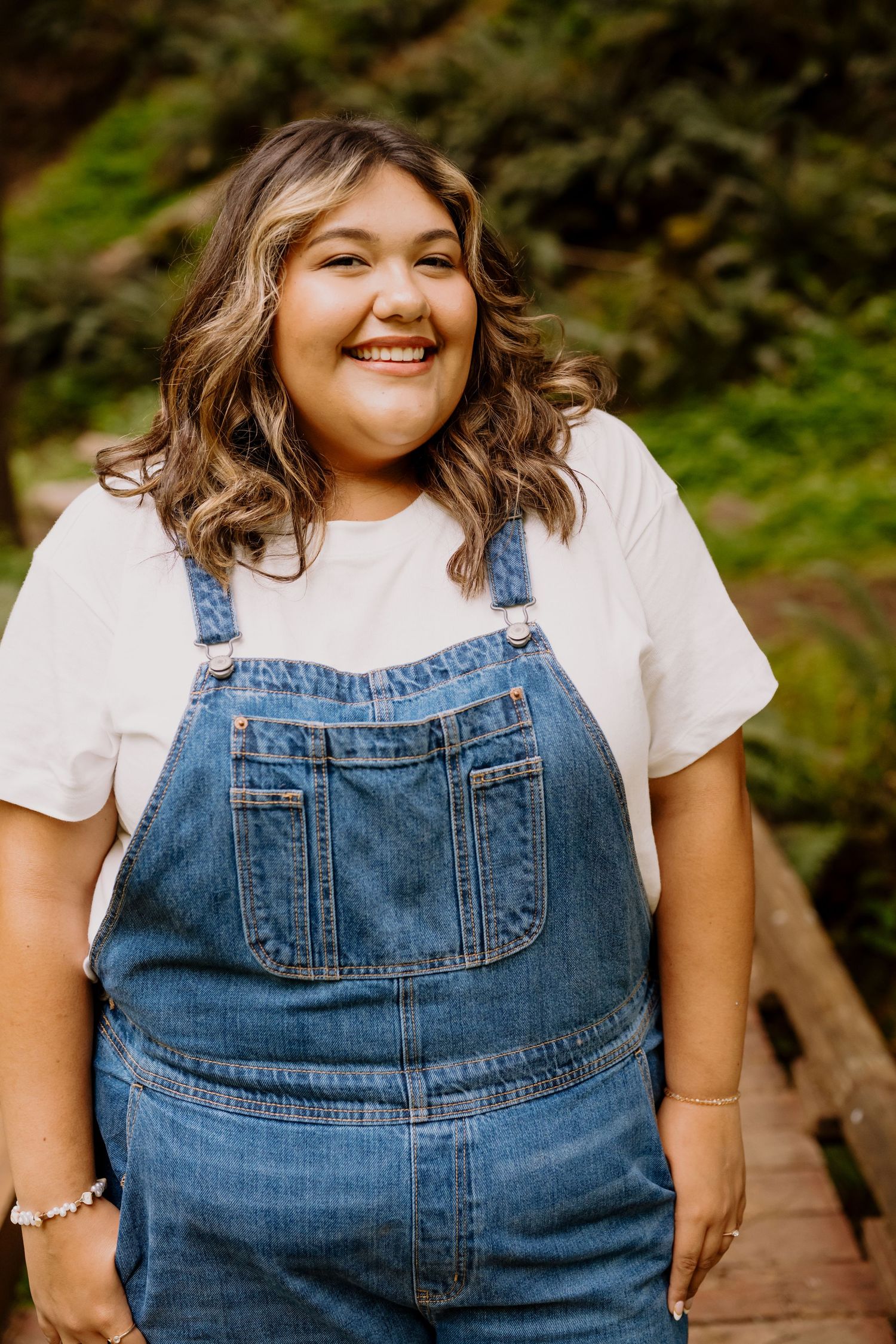 Smiling young woman in denim overalls and white t-shirt posing outdoors on a wooden bridge.