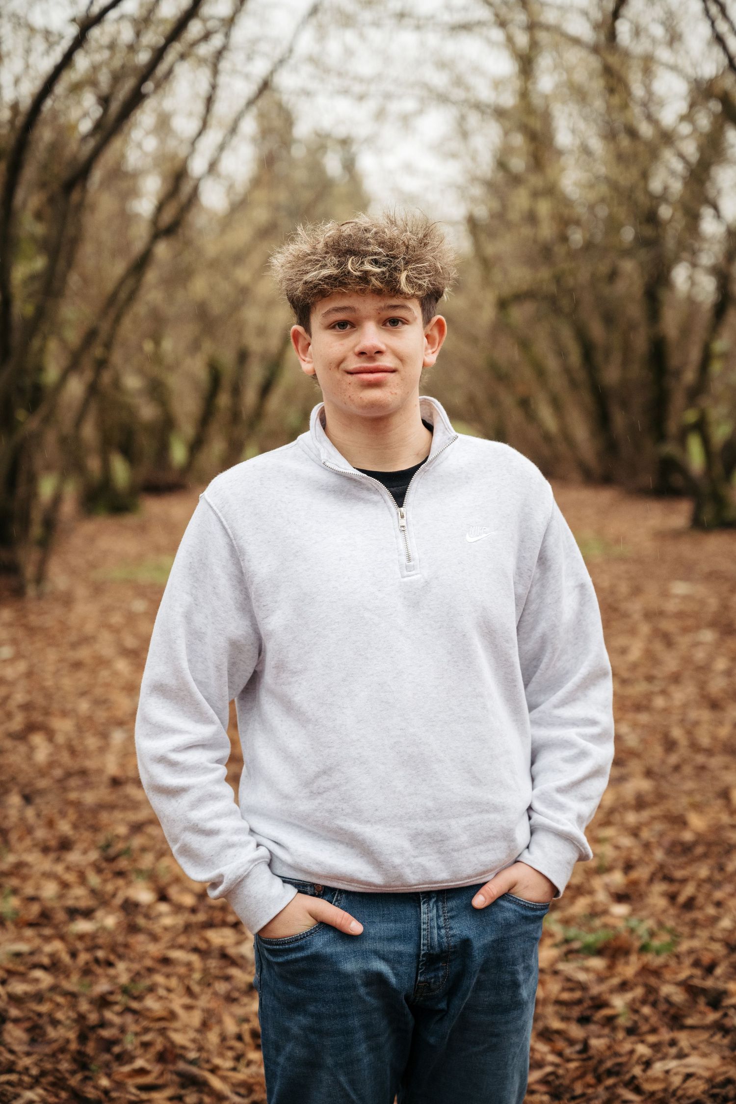 Person in white quarter-zip sweater and jeans poses outdoors among fallen autumn leaves.
