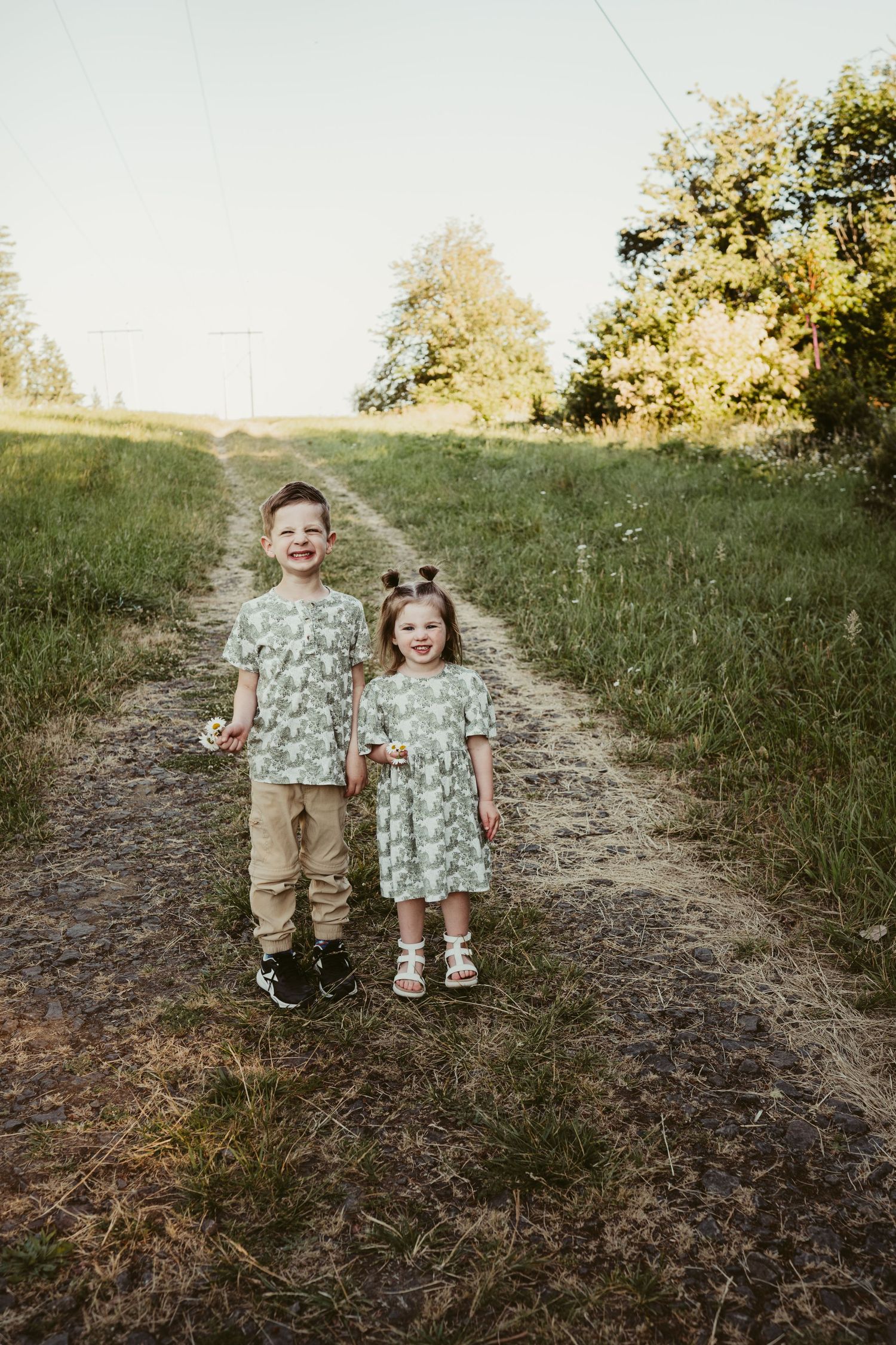 Two young siblings stand together on a dirt path through a grassy hillside during golden hour.